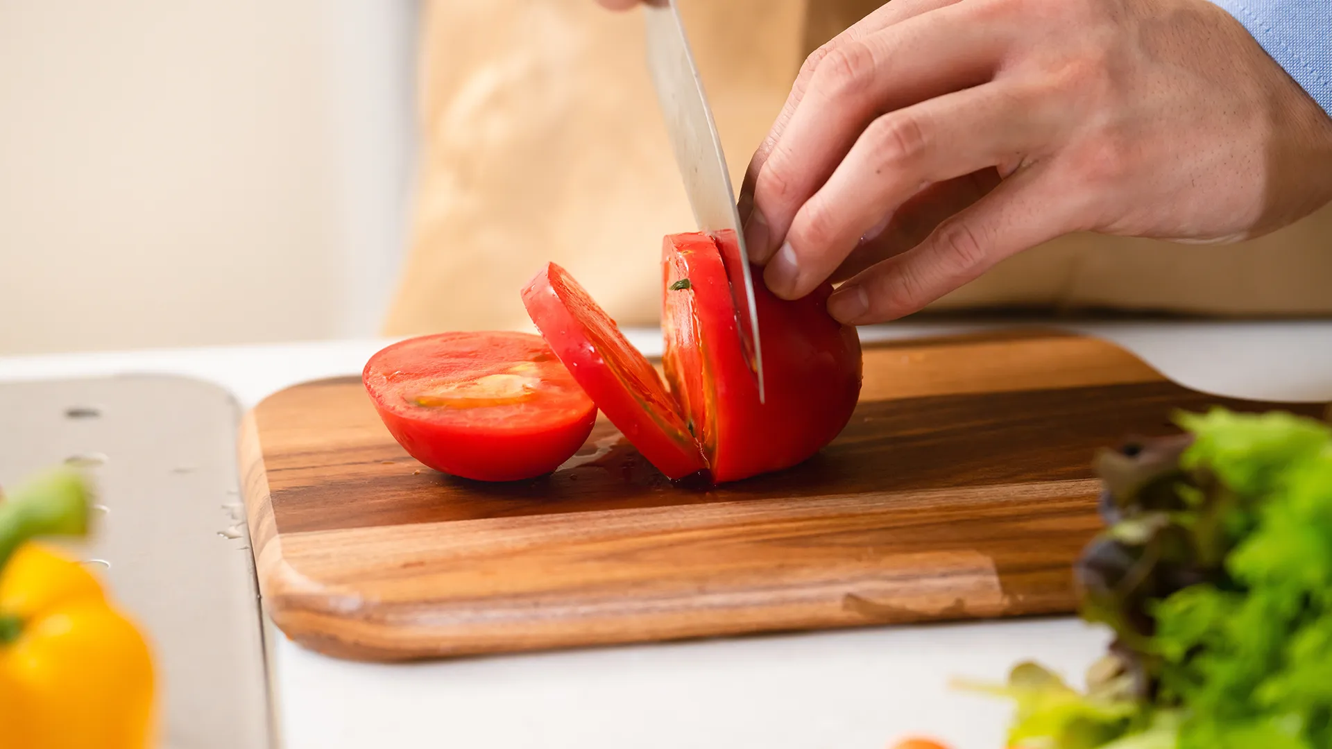 A hand holds down a tomato while the other uses a sharp knife to slice it on a wooden chopping board. On the foreground some other vegetables are visible. 