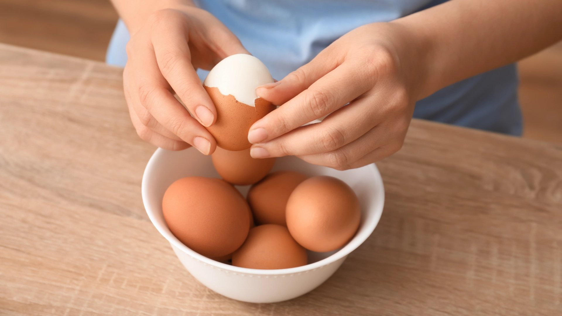 Two hands de-shelling a hard boiled egg over a bowl containing five eggs.