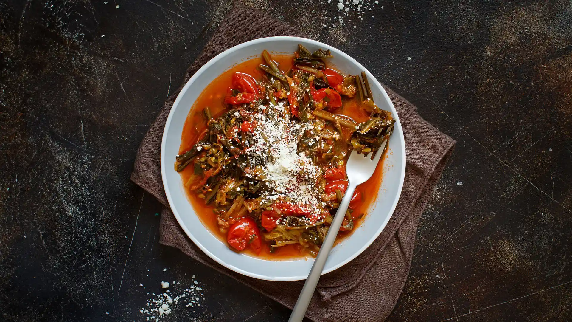 A top-down view of a white plate with Ribollita; featuring pieces of cooked vegetables and soaked bread pieces on top of a tomato sauce. 