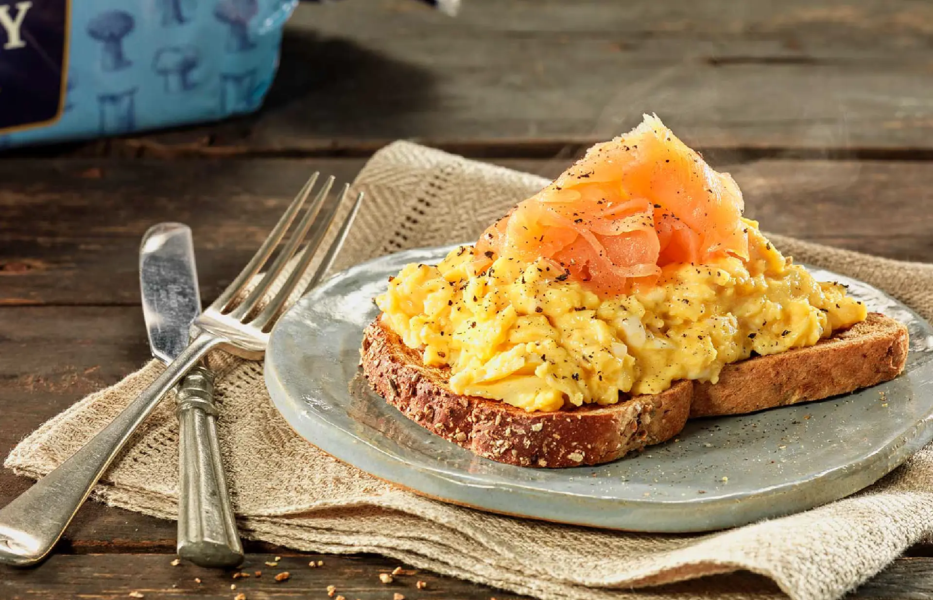 A slice of Hovis Granary Medium Sliced on a plate, topped with scrambled eggs, slices of smoked salmon, and a sprinkle of black pepper.