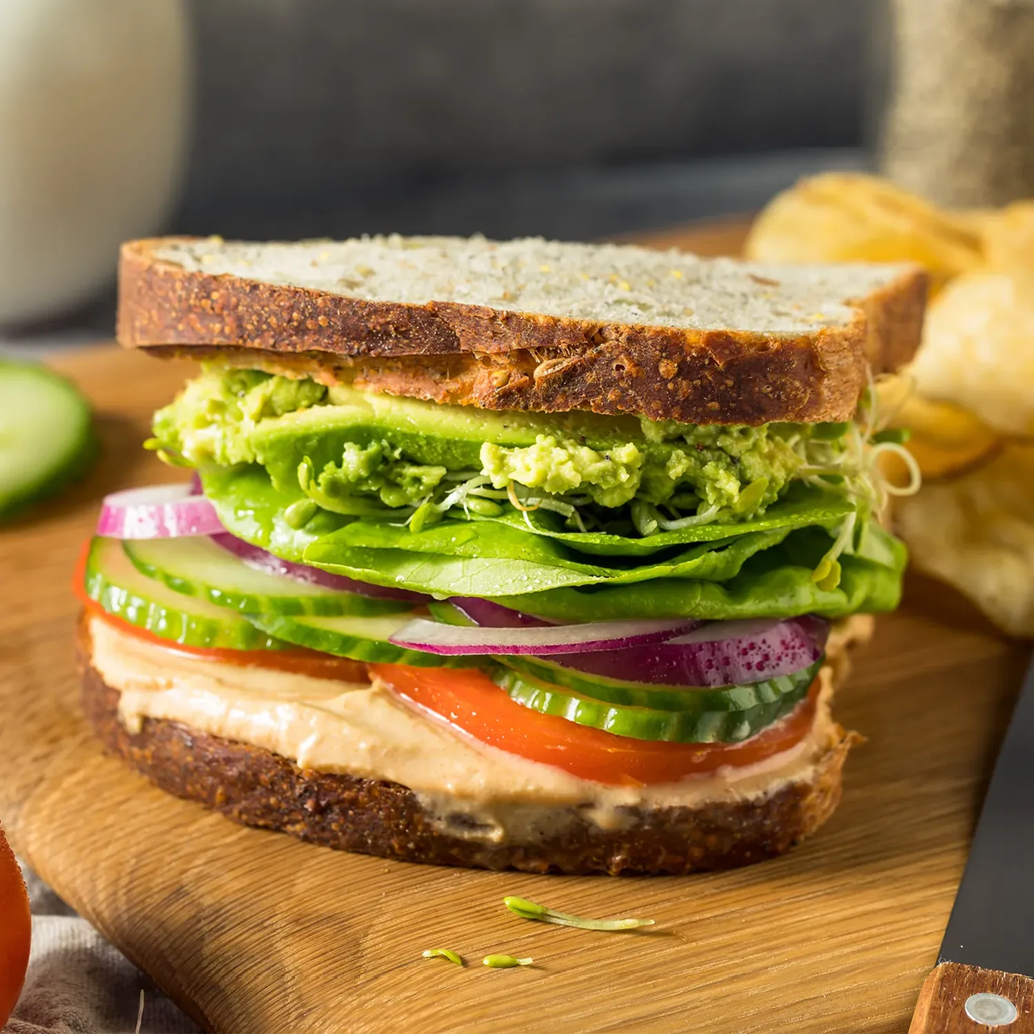 A Hovis Tasty Wholemeal veggie sandwich rests on a wooden table, filled with sliced tomatoes, red onions, mashed avocado, lettuce and hummus.