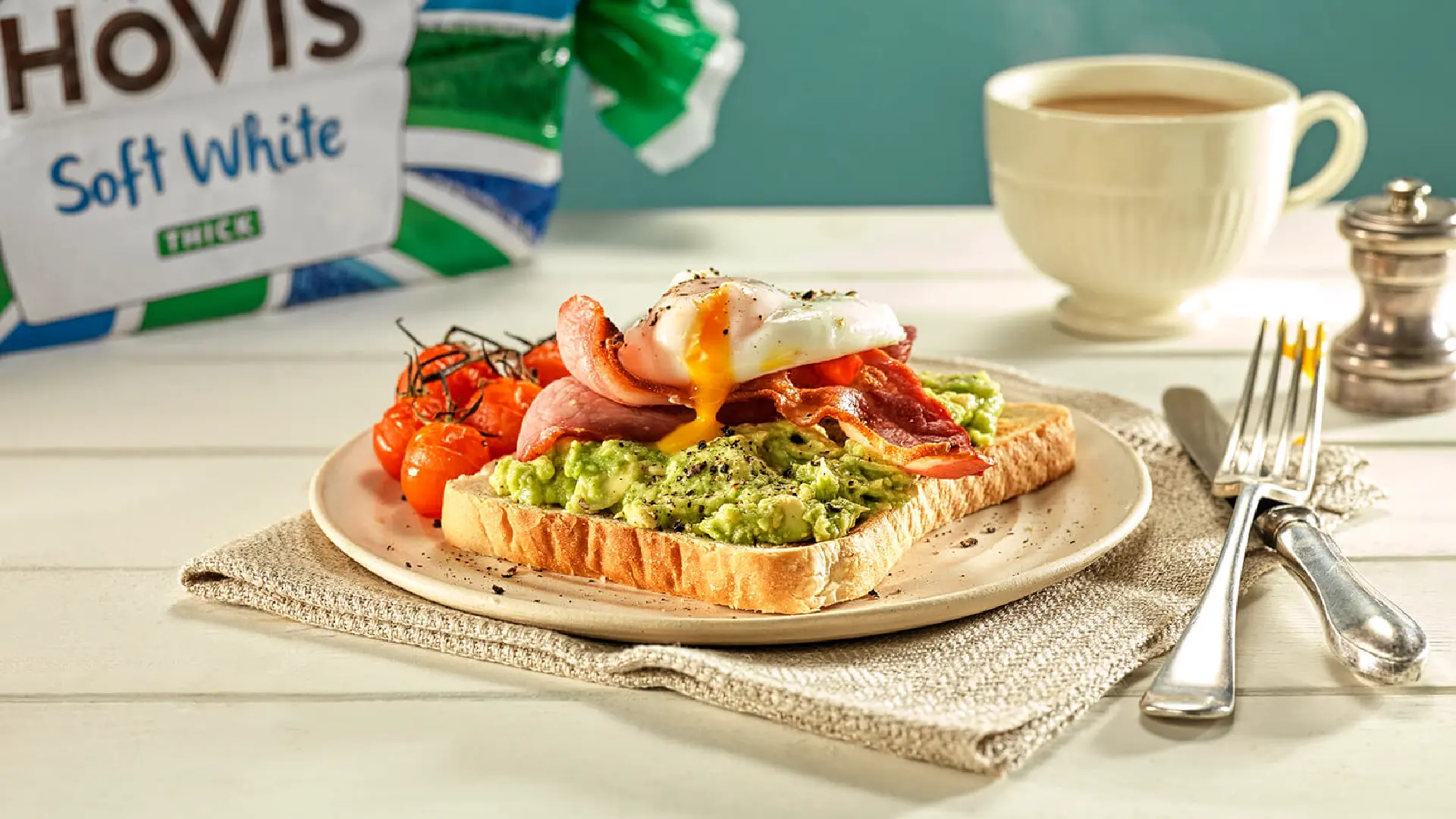 A slice of white bread on a plate, topped with mashed avocado, crispy bacon, a poached egg with yellow yolk running and some grilled vine cherry tomatoes on the side. 