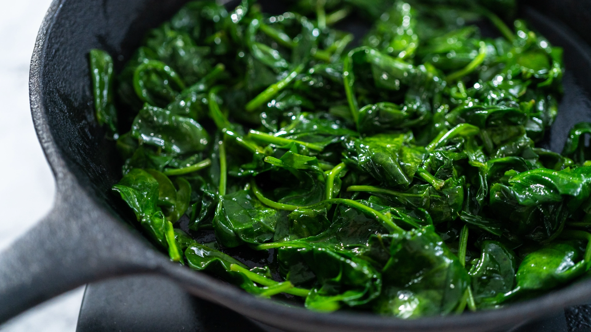 A black iron frying pan sits on the hob with cooked green spinach leaves. 