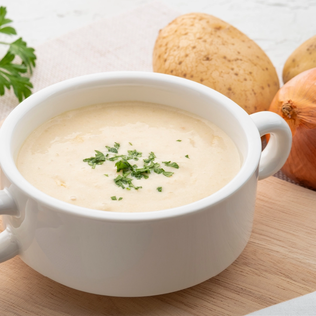 A white bowl filled with creamy beige potato soup, topped with sliced parsley, sits on a wooden chopping board. In the background are two potatoes, an onion, and some parsley leaves.