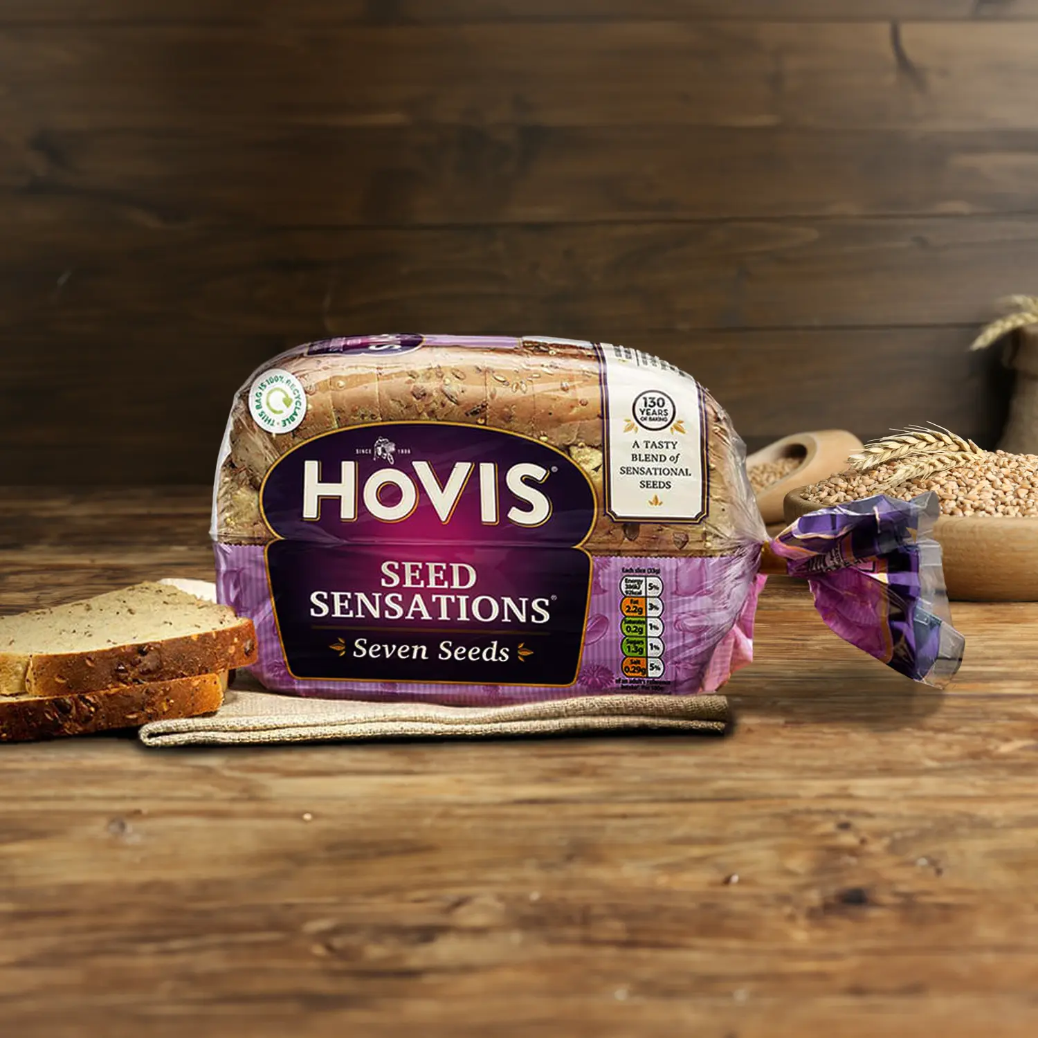 Hovis Seed Sensations Original 400g on a wooden table, with a bowl of wheat grains, a wooden scoop, and dried wheat in the background, along with slices of bread on the left.