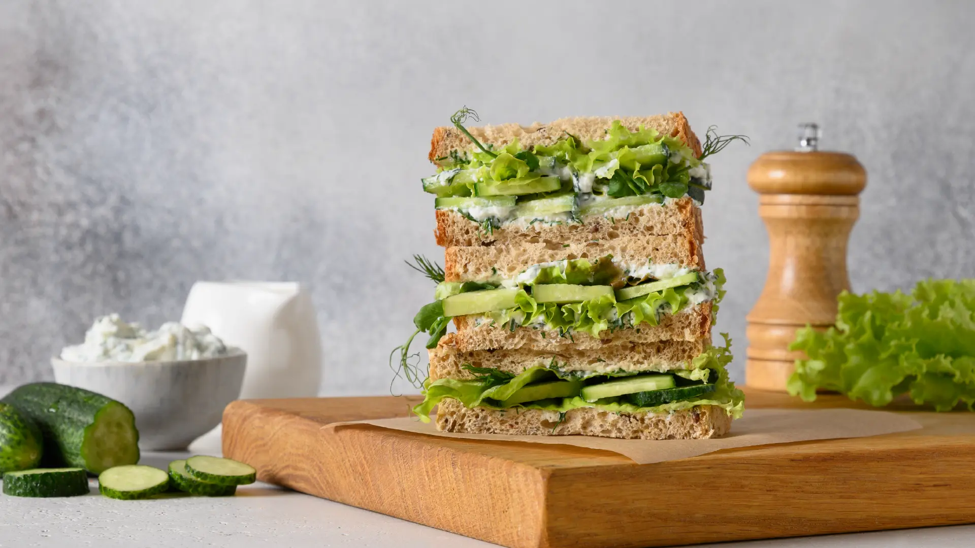 Three sandwiches stacked on a wooden board, surrounded by a small bowl of cream cheese and sliced cucumber. Salad leaves, cucumber slices, cream cheese, and dill are spilling out from between the bread slices.