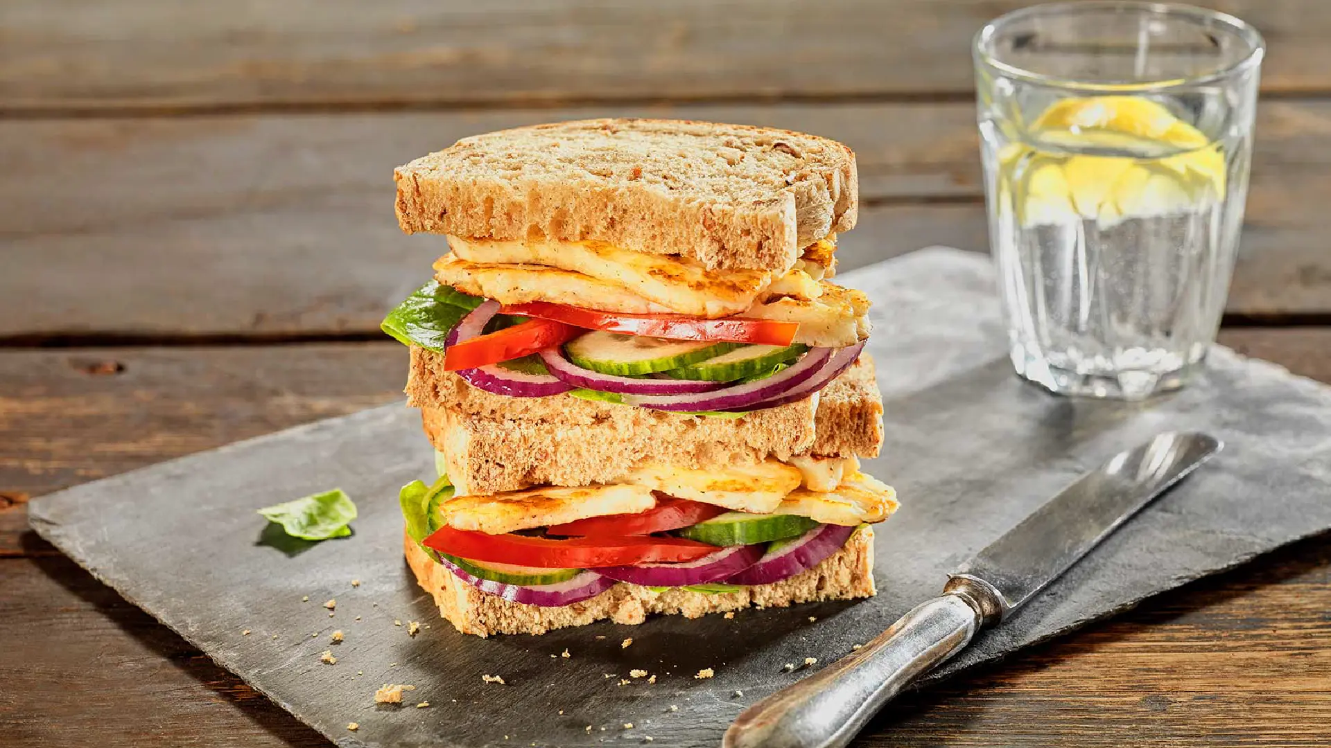 Two sandwiches stacked, filled with slices of red onion, cucumber, red pepper, and halloumi between brown bread slices.