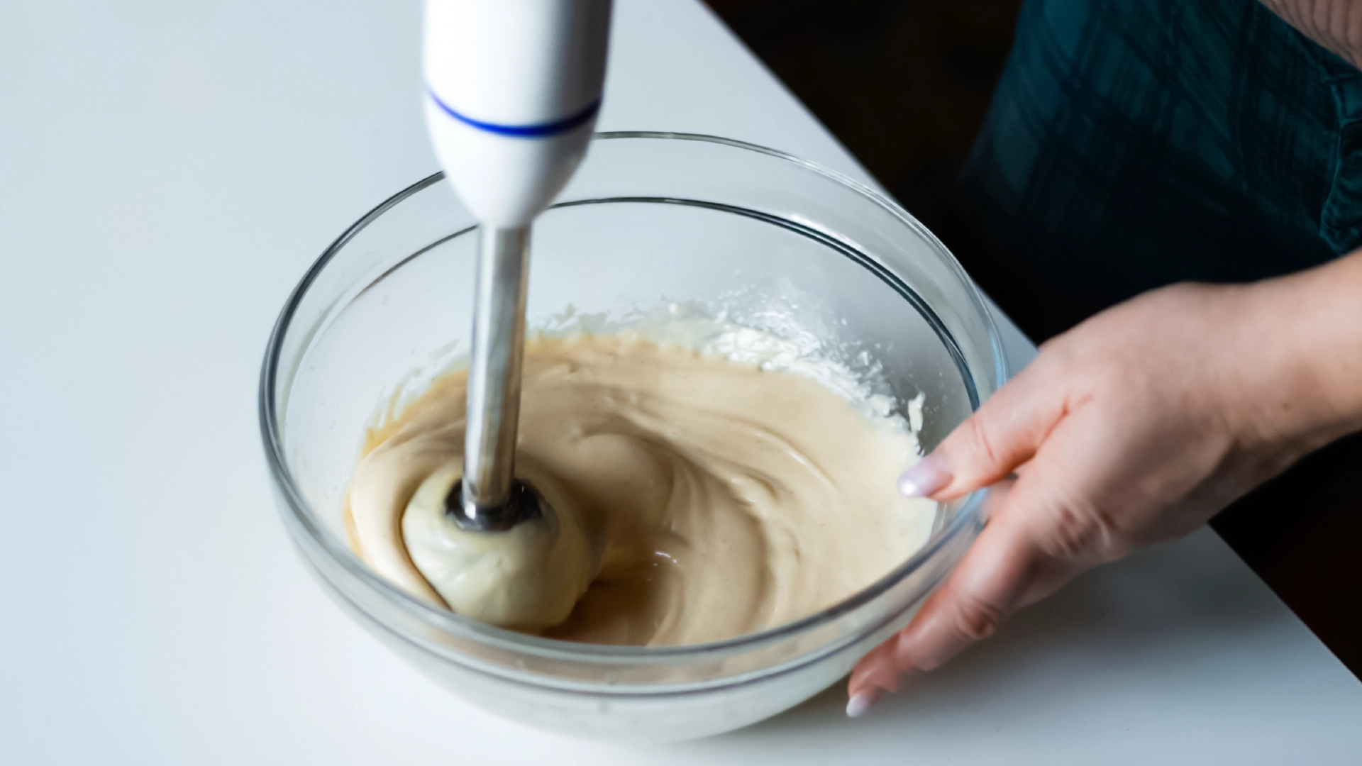 One hand holding a glass bowl while the other uses a hand blender to mix the mayonnaise.