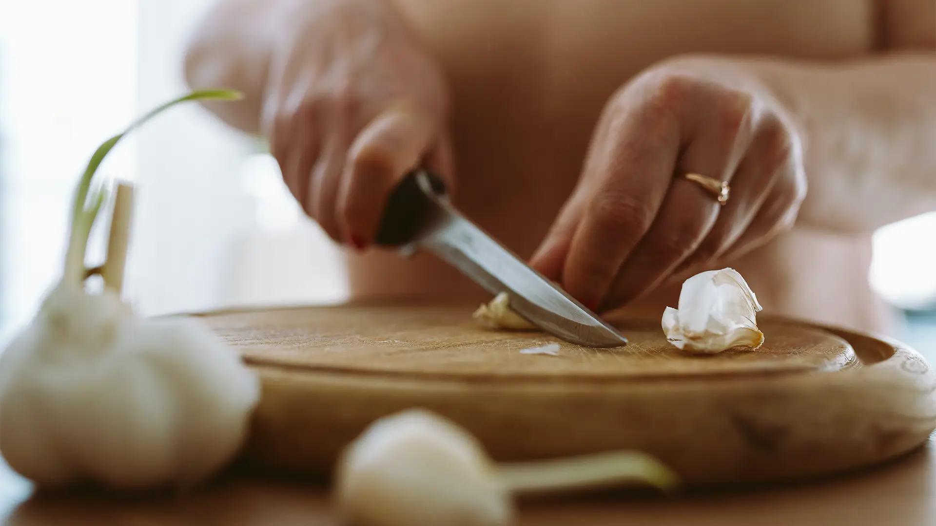 A hand holds down a garlic clove, while the other uses a sharp knife to cut a small piece off on a round, wooden, chopping board. Some extra garlic can be seen on the foreground. 