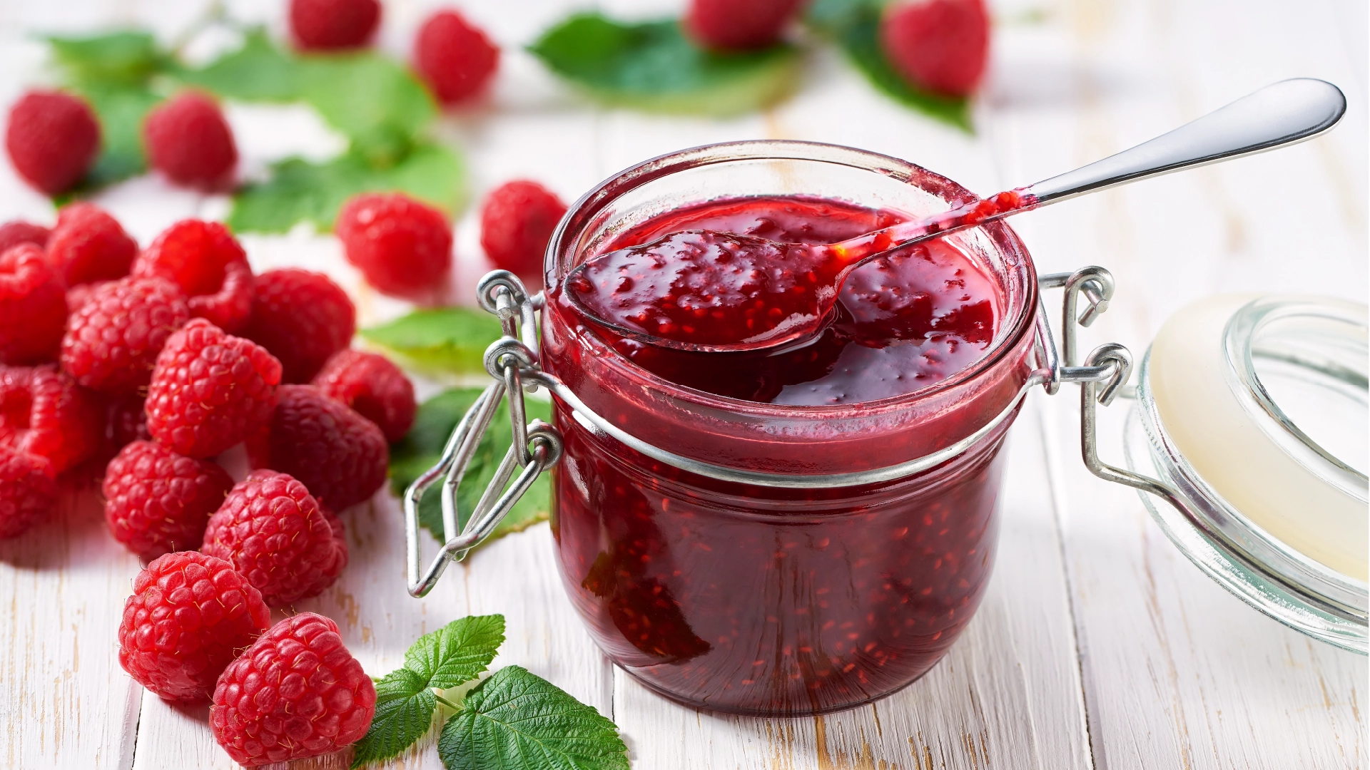 A glass jar filled with raspberry jam, with a spoon full of jam resting on top, surrounded by fresh raspberries and mint leaves.