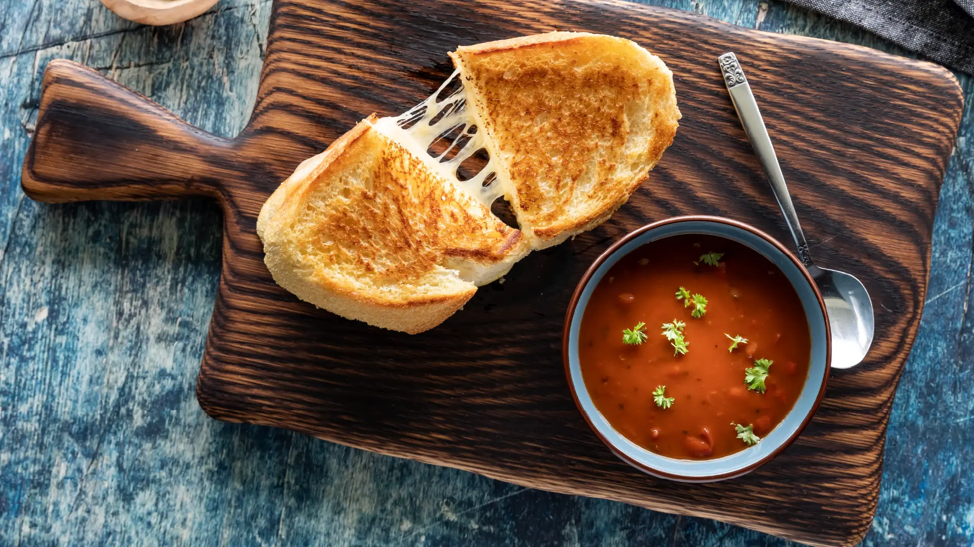 A top-down view of a bowl filled with tomato soup garnished with green herbs, alongside a sliced cheese toastie.