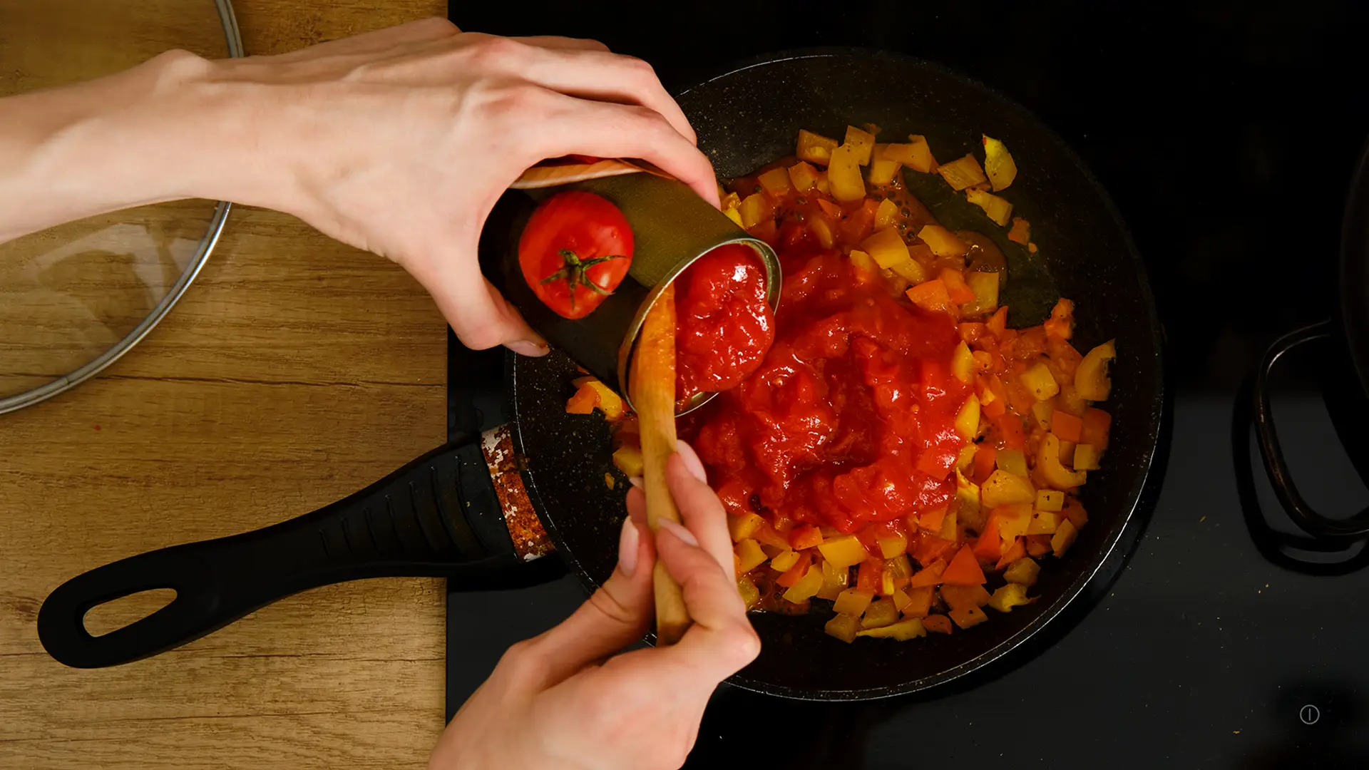 Someone is using a wooden spoon to pour a tin of chopped tomatoes into a black metal pan filled with sautéed vegetables.