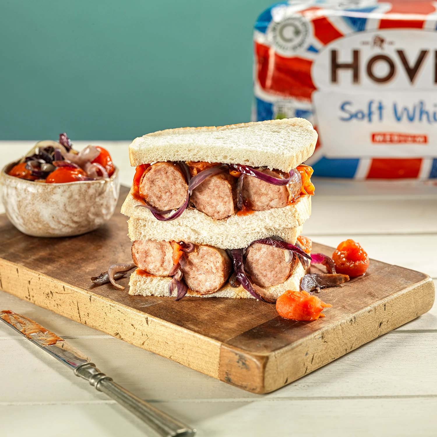 Two halves of a sandwich made with Hovis Soft White Medium Sliced bread, stacked and filled with red onions, sausage, and roasted tomatoes, on a wooden chopping board.