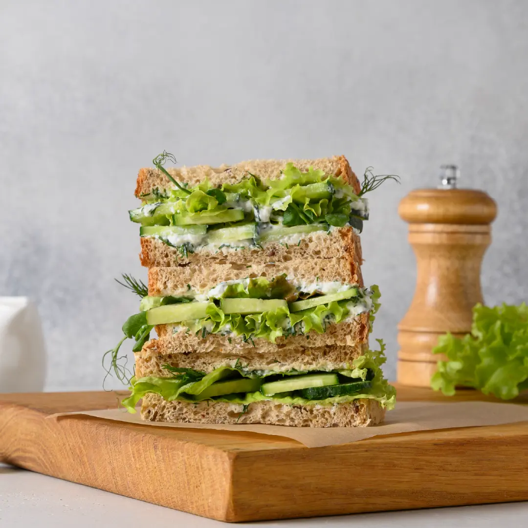 Three sandwiches stacked on a wooden board, surrounded by a small bowl of cream cheese and sliced cucumber. Salad leaves, cucumber slices, cream cheese, and dill are spilling out from between the bread slices.