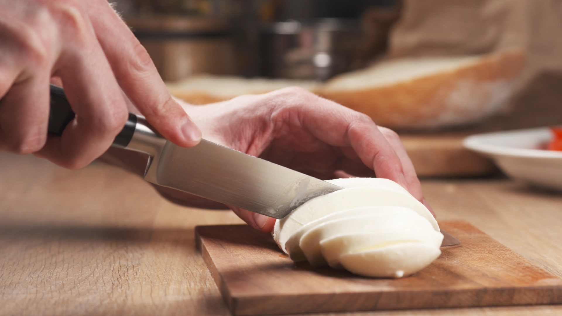 A hand holds a mozzarella ball while the other uses a sharp knife to slice it into thinner pieces.