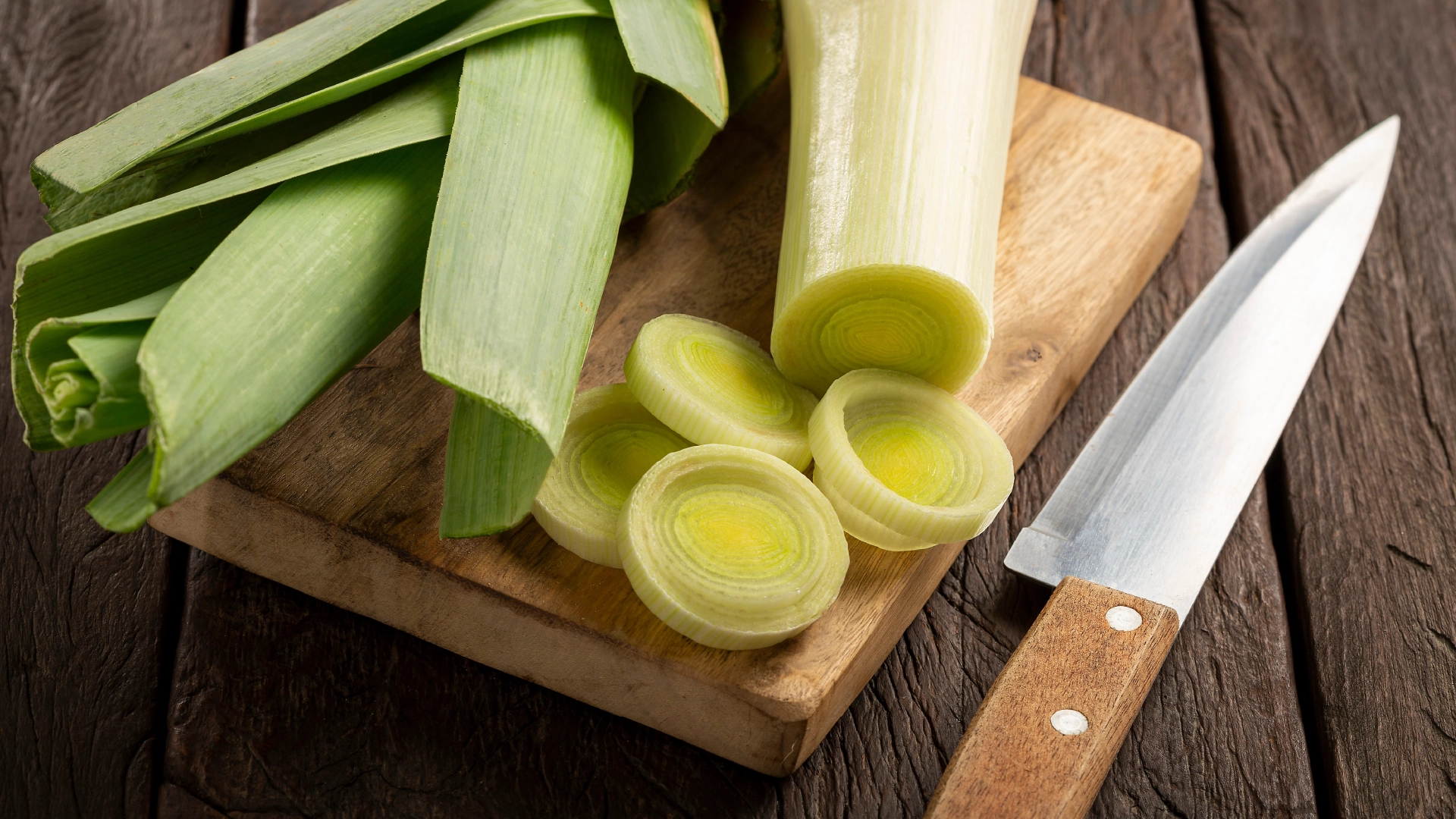 A leek next to four round slices on a wooden board, with a sharp knife on the right.