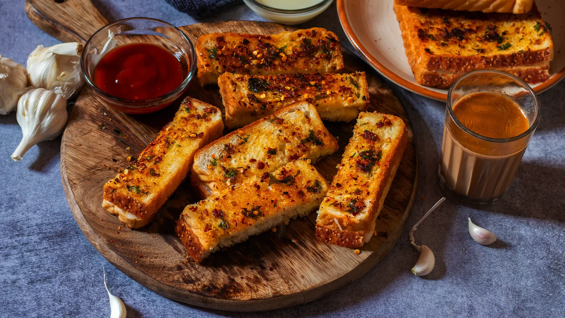 Hovis Seed Sensations Original bread, sliced in half and placed on a wooden chopping board. The slices are golden brown, topped with melted cheese and a sprinkle of thinly sliced herbs. On the side, a small bowl of ketchup and a few garlic cloves.