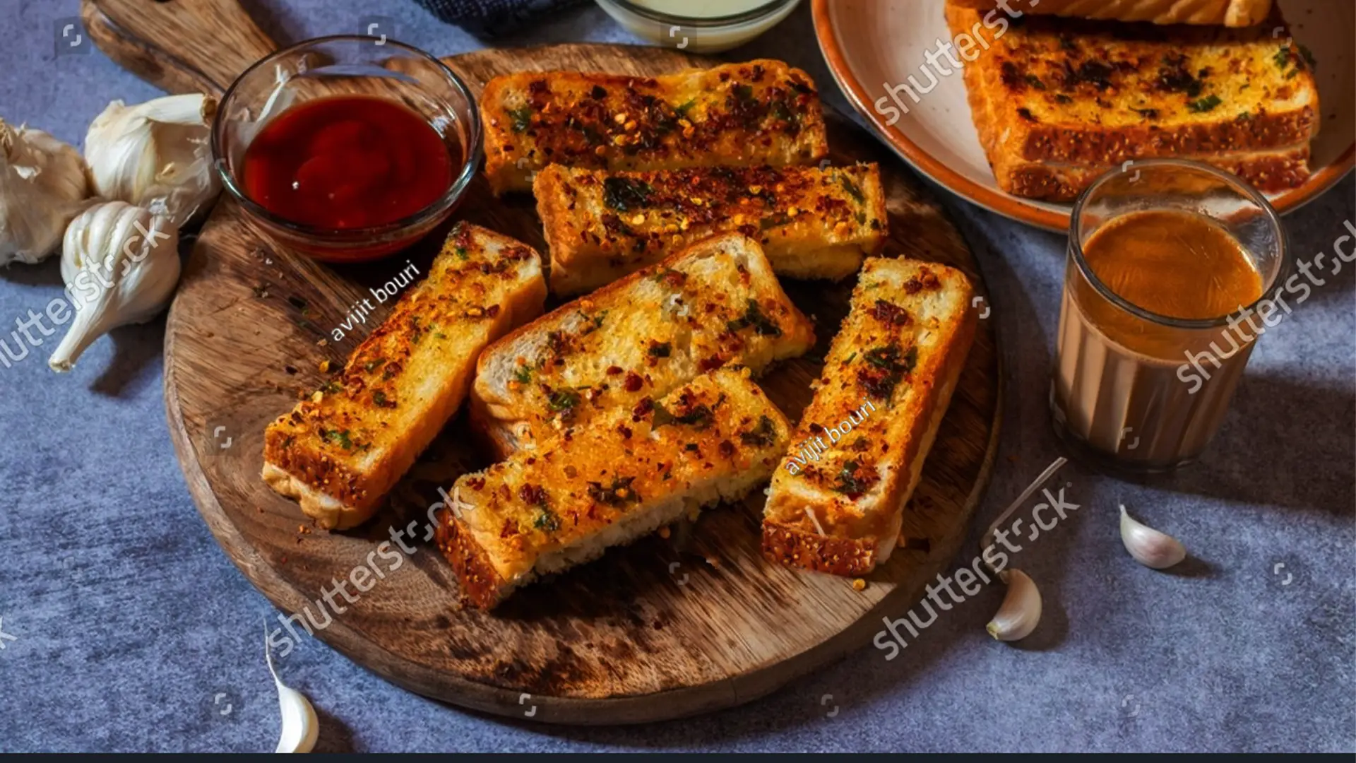 Hovis Seed Sensations Original bread, sliced in half and placed on a wooden chopping board. The slices are golden brown, topped with melted cheese and a sprinkle of thinly sliced herbs. On the side, a small bowl of ketchup and a few garlic cloves.