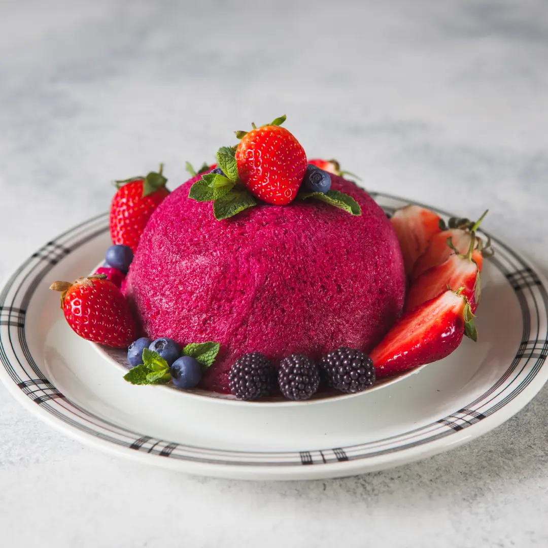 A small, bright pink dome-shaped pudding on a plate, topped with a strawberry and mint leaves. The base is surrounded by halved strawberries, blackberries, blueberries, and more mint leaves.