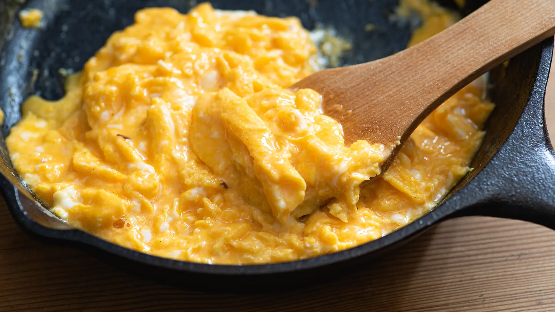 Creamy and silky scrambled eggs in a black frying pan with a wooden spatula.