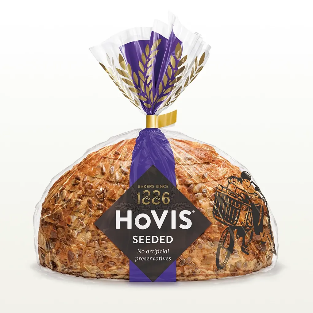 Hovis Seeded Half Cob bread on a white backdrop with a subtle shadow, featuring packaging decorated with a dark purple stripe and golden wheats.