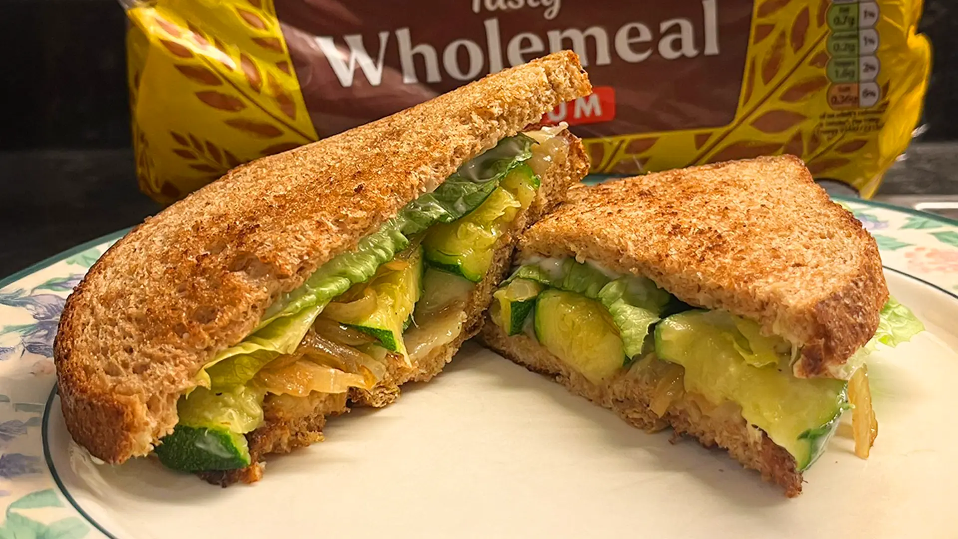 A toasted wholemeal sandwich with lettuce and courgette, cut in half and held by two hands. A loaf of wholemeal bread is in the background.