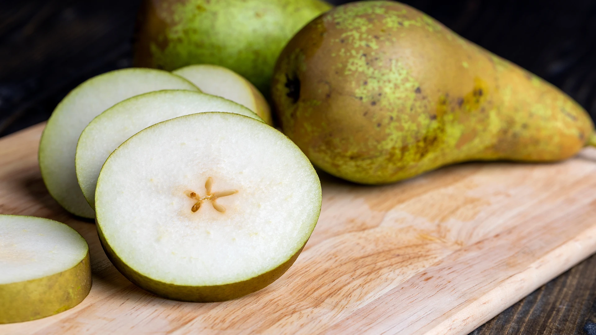 Hands using a small, sharp knife to cut a quarter of a pear into thin slices on a wooden board.