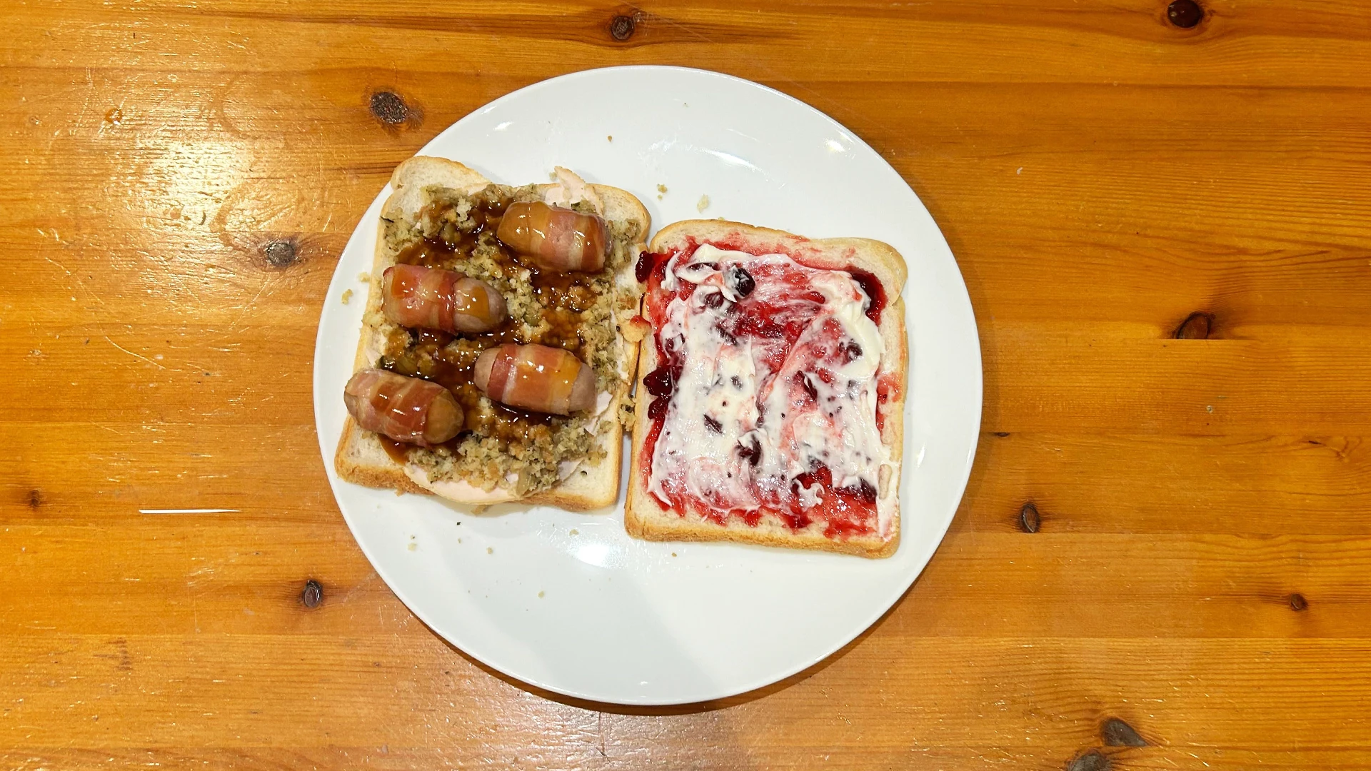 A white plate with two pieces of Hovis Soft White Bread, on one slice it has stuffing, turkey and pigs in blankets with some gravy. On the other slice is cranberry and mayonnaise.