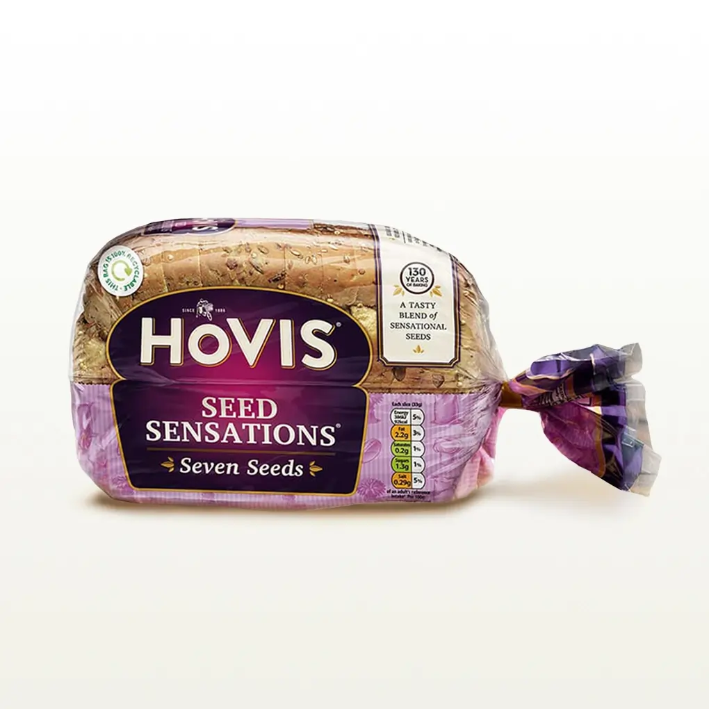 Hovis Seed Sensations Original 400g bread on a white backdrop with a subtle shadow. The packaging is half transparent and half pink, decorated with images of flour, a chefs hat, seeds and wheat.