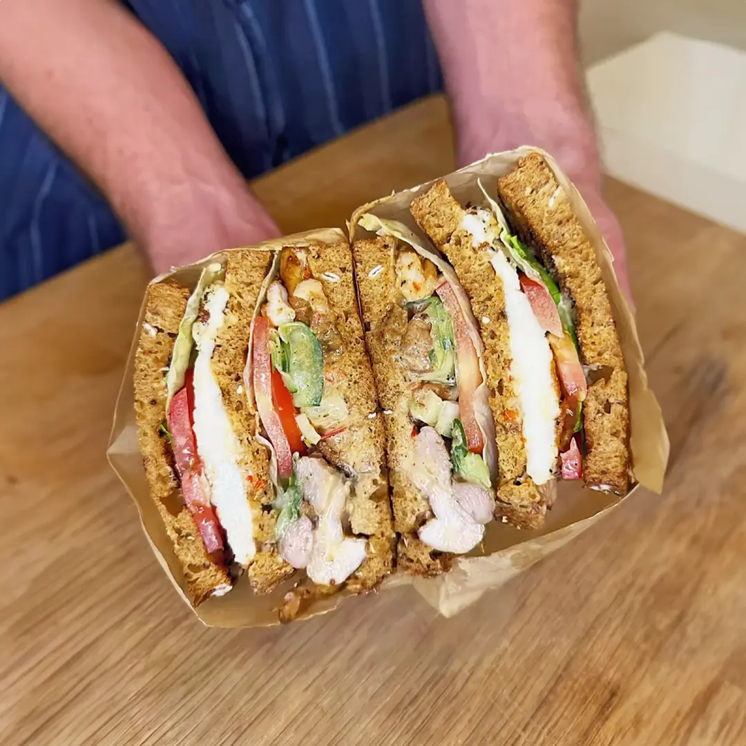 Someone holding two halves of a sandwich wrapped in paper. The sandwich is made of brown bread, filled with tomato slices, lettuce, juicy grilled chicken, and thin cucumber slices.