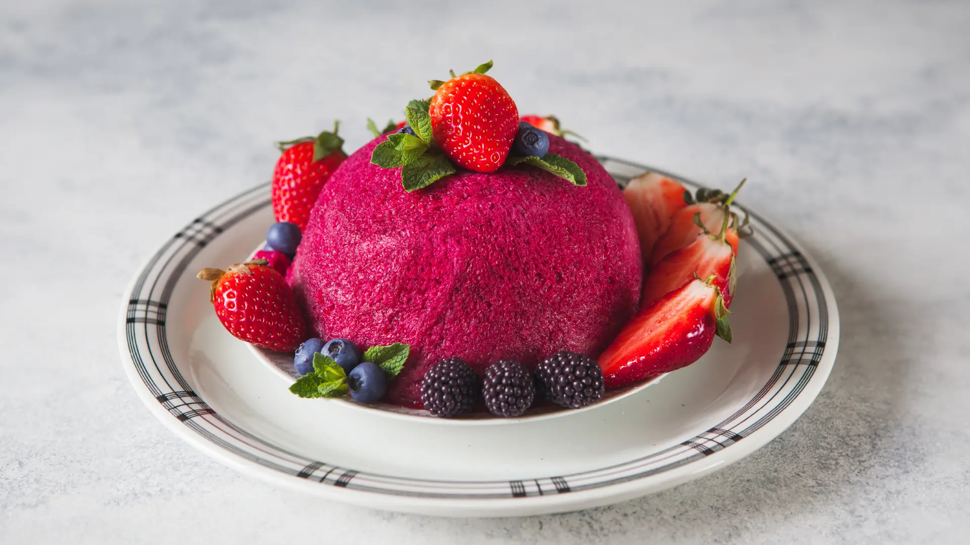 A small, bright pink dome-shaped pudding on a plate, topped with a strawberry and mint leaves. The base is surrounded by halved strawberries, blackberries, blueberries, and more mint leaves.