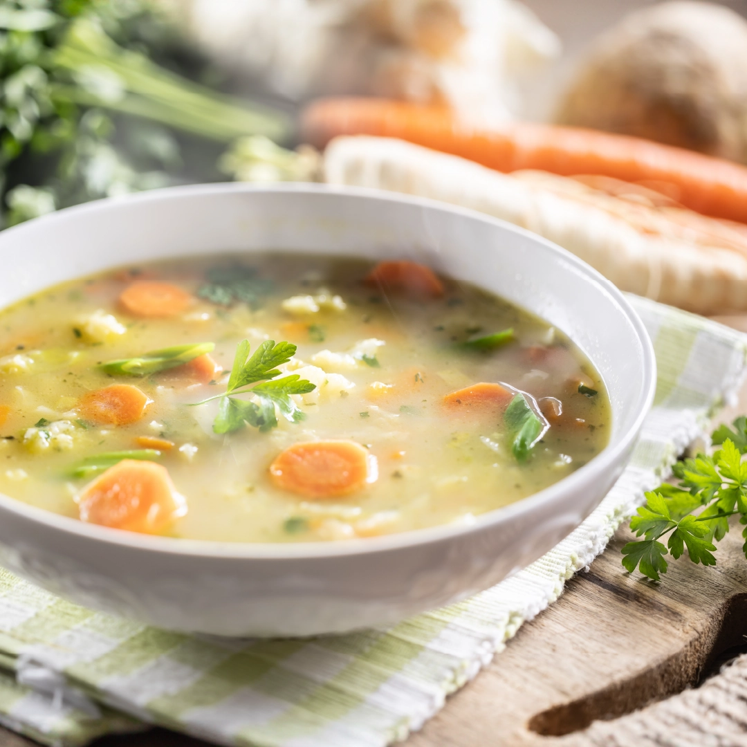 A bowl of chicken soup with steam rising, featuring round slices of carrot, chopped greens, and a parsley leaf on top.