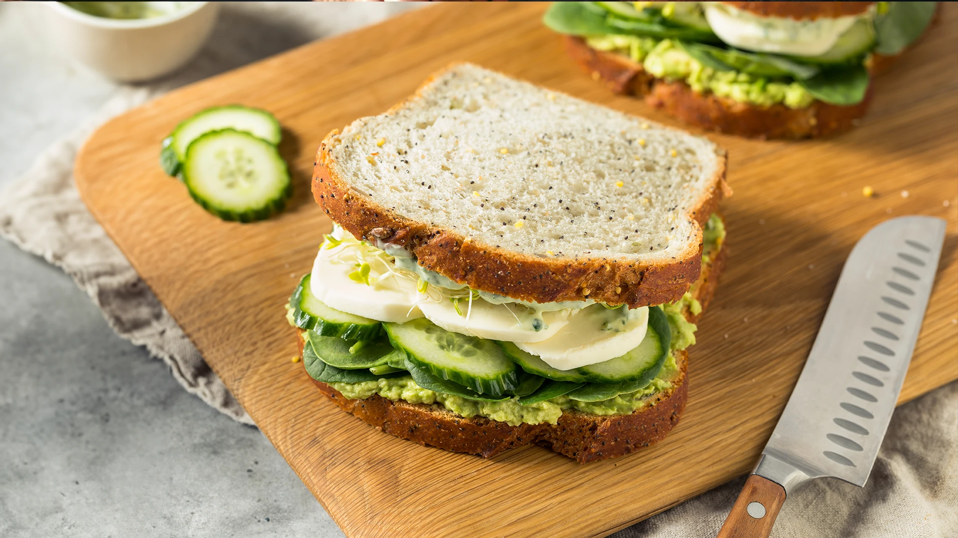 A seeded bread sandwich filled with mashed avocado, sliced cucumber, and mozzarella cheese, with cucumber slices and a knife on the side.