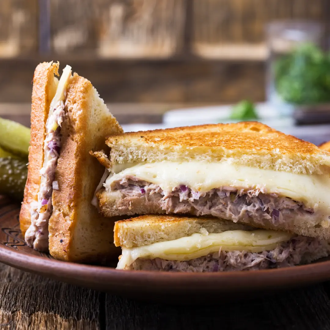 Four slices of toasted brown bread sandwiches on a plate, filled with tuna mayonnaise and molten cheese oozing between the slices.