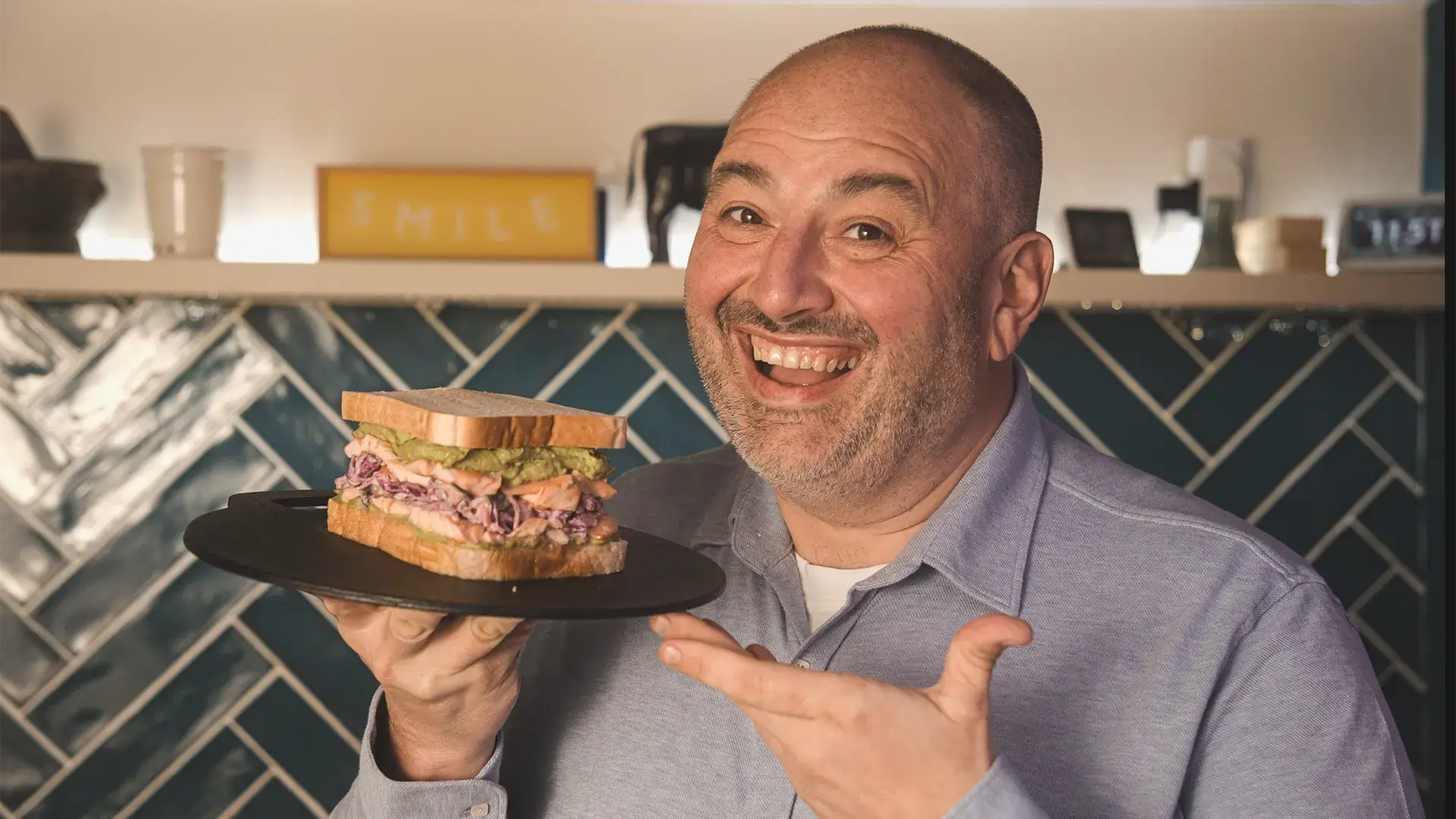 Wynne Evans holds up his Sushi Sandwich on a plate and smiles happy at the camera. Behind him is his kitchen.