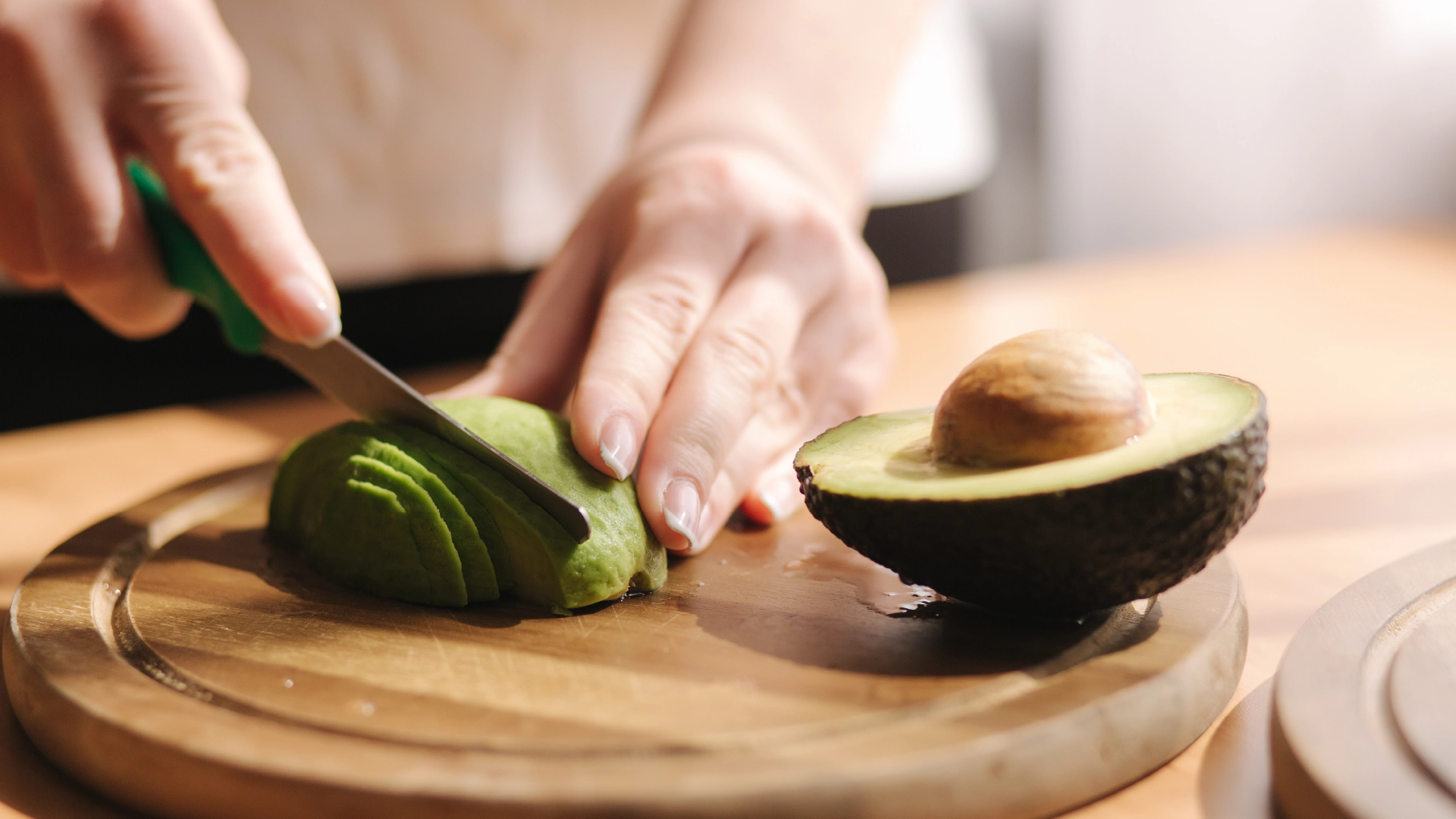 A set of hands slicing one half of an avocado lengthwise on a wooden chopping board.