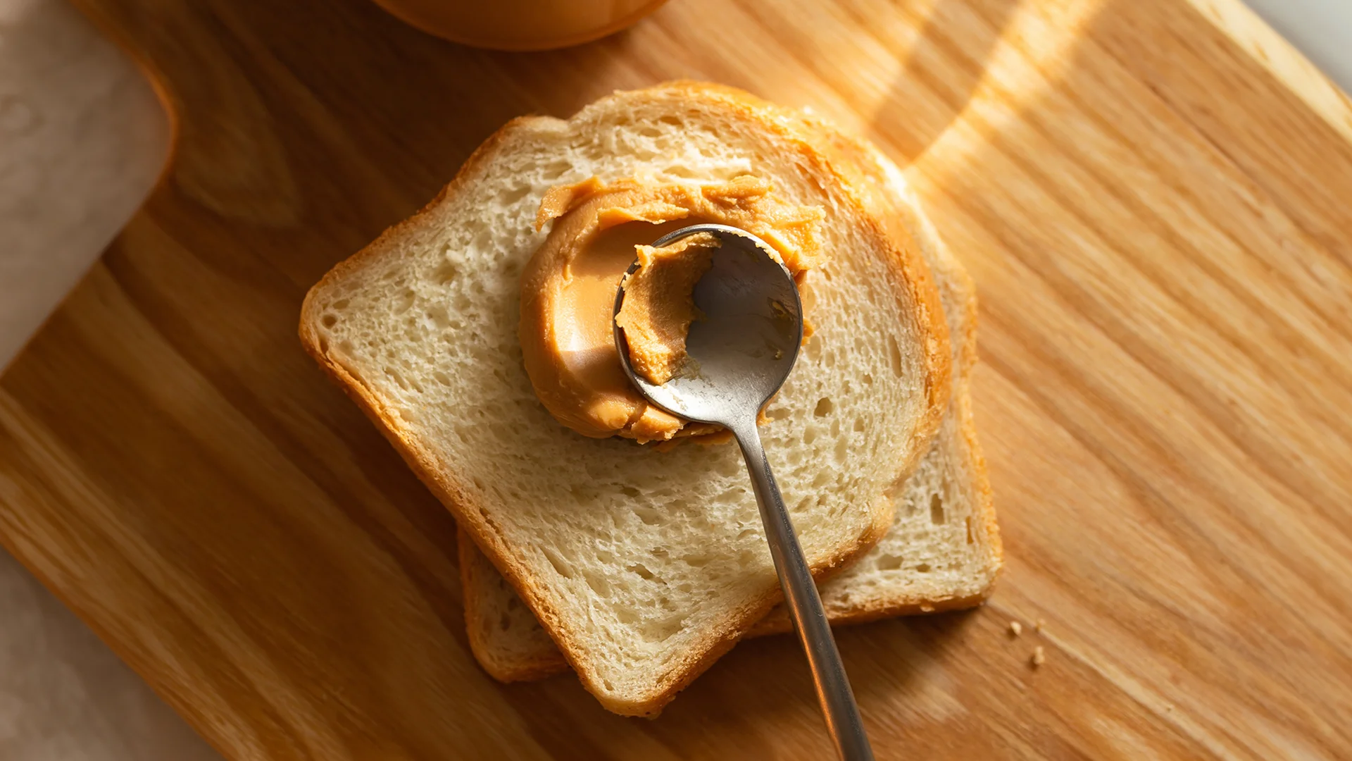 Hovis® Soft White bread on a wooden chopping board, with smooth peanut butter being spread onto it with a spoon.