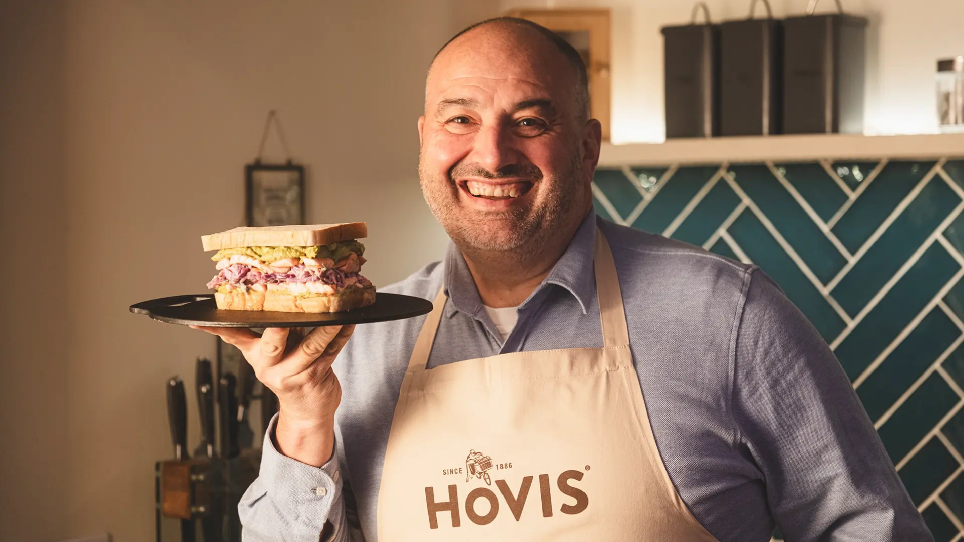 A man in a Hovis apron holds a plate with a sandwich filled with mashed avocado, salmon, and coleslaw.