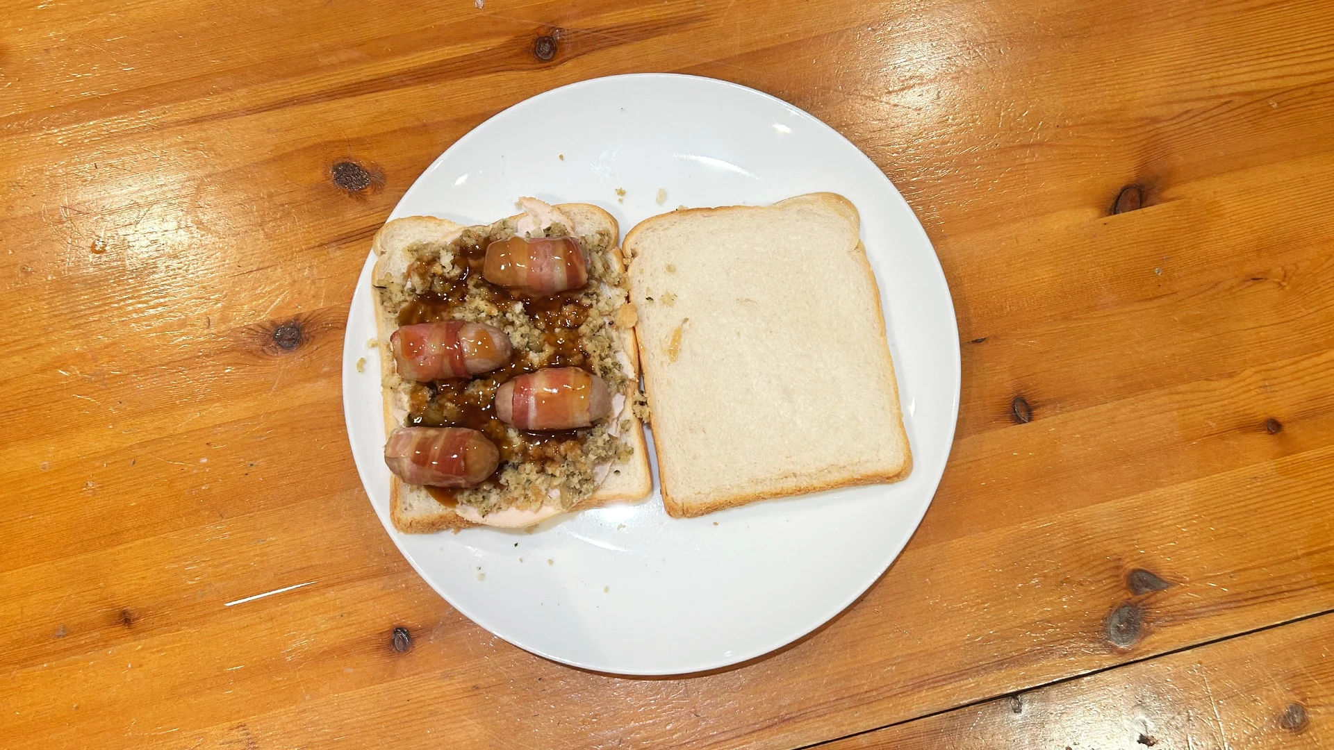  Hovis Soft White Medium Sliced bread topped with turkey slices, stuffing, and crispy pigs in blankets, placed on a plate on a kitchen counter.