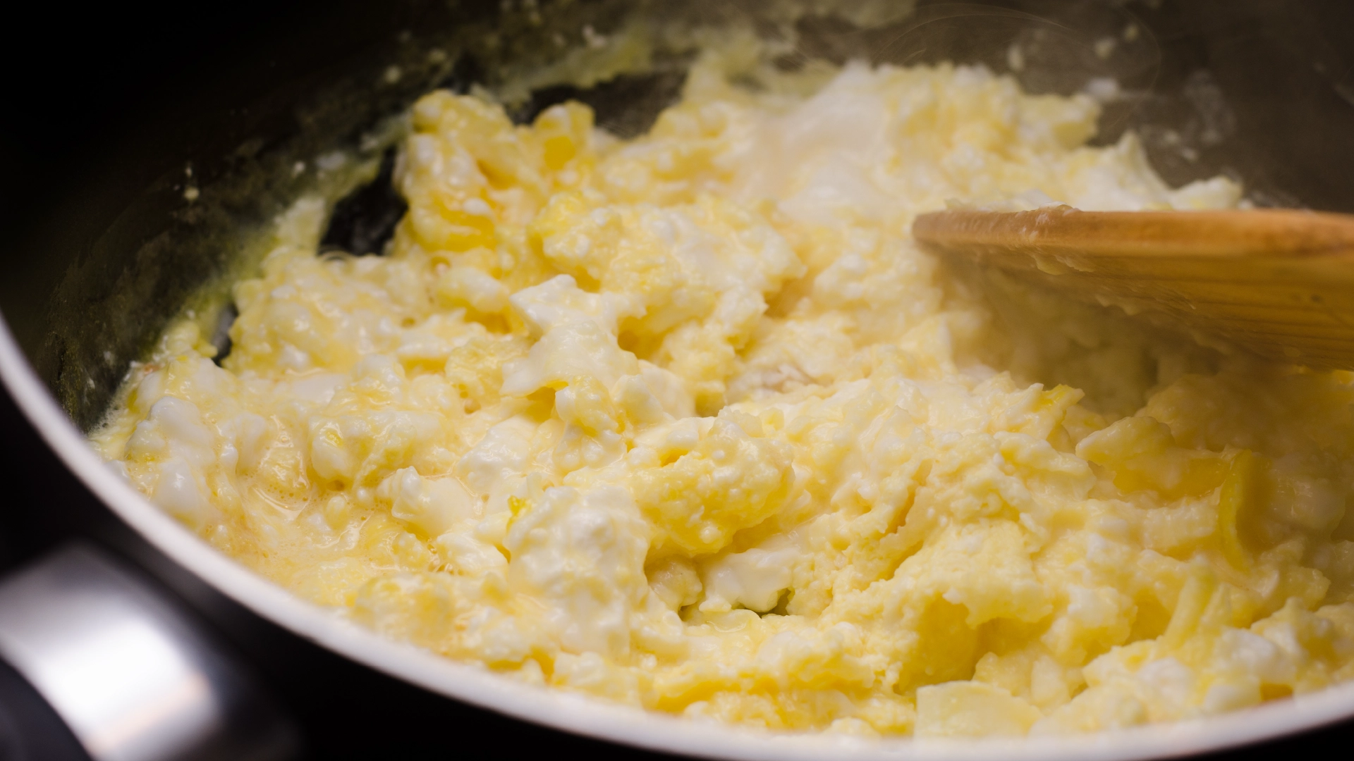 A frying pan sits on the hob with cooked scrambled eggs. A wooden spatula mixes the eggs, folding them gently to ensure even cooking.