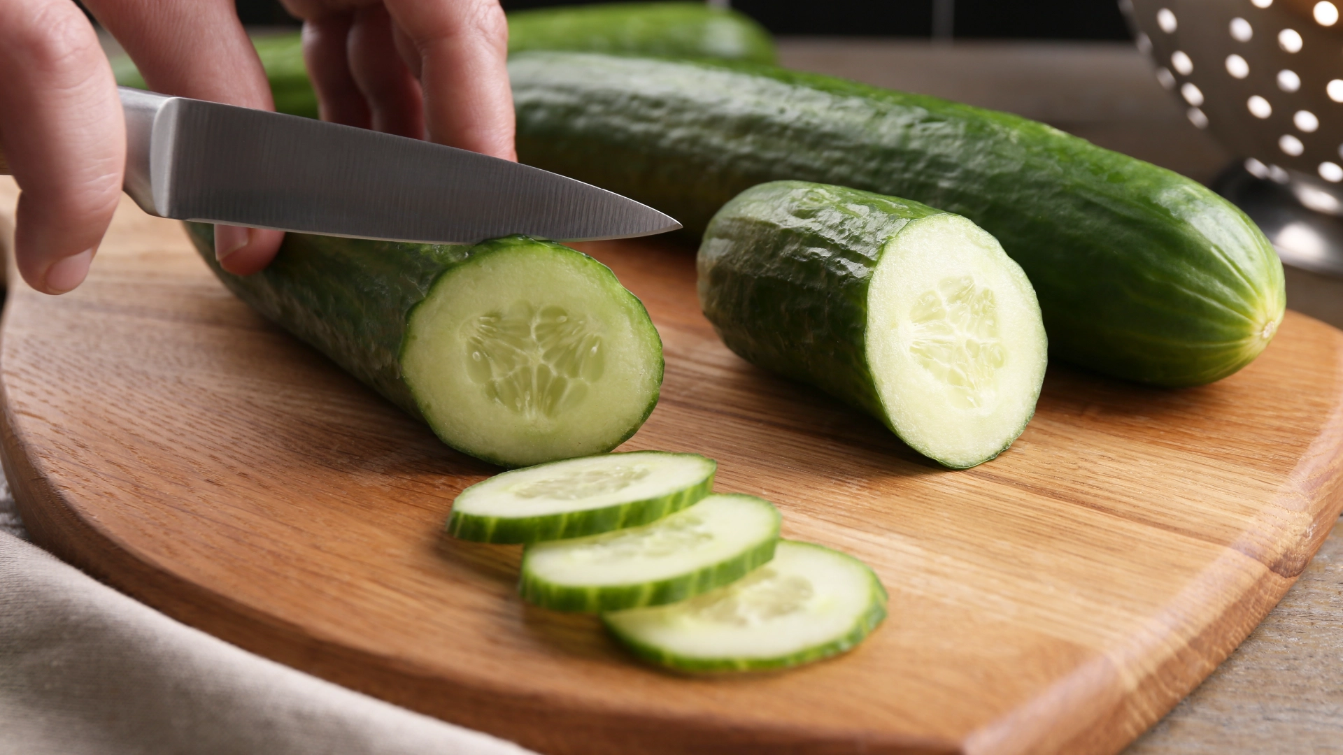 Hands using a small, sharp knife to thinly slice a cucumber on a wooden chopping board.