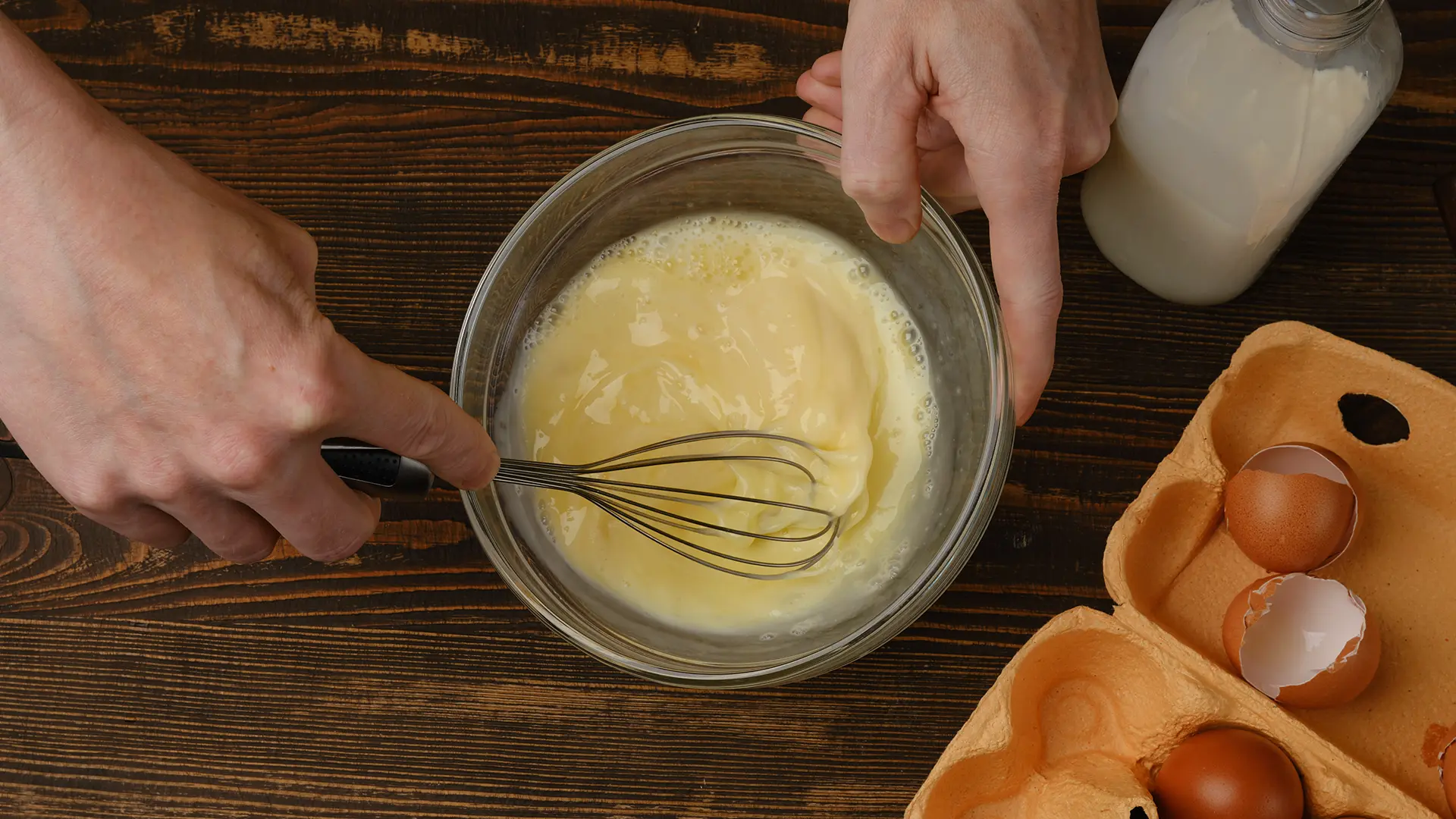 A slurry of milk and tapioca starch being whisked into some eggs in a glass bowl on a wooden countertop.