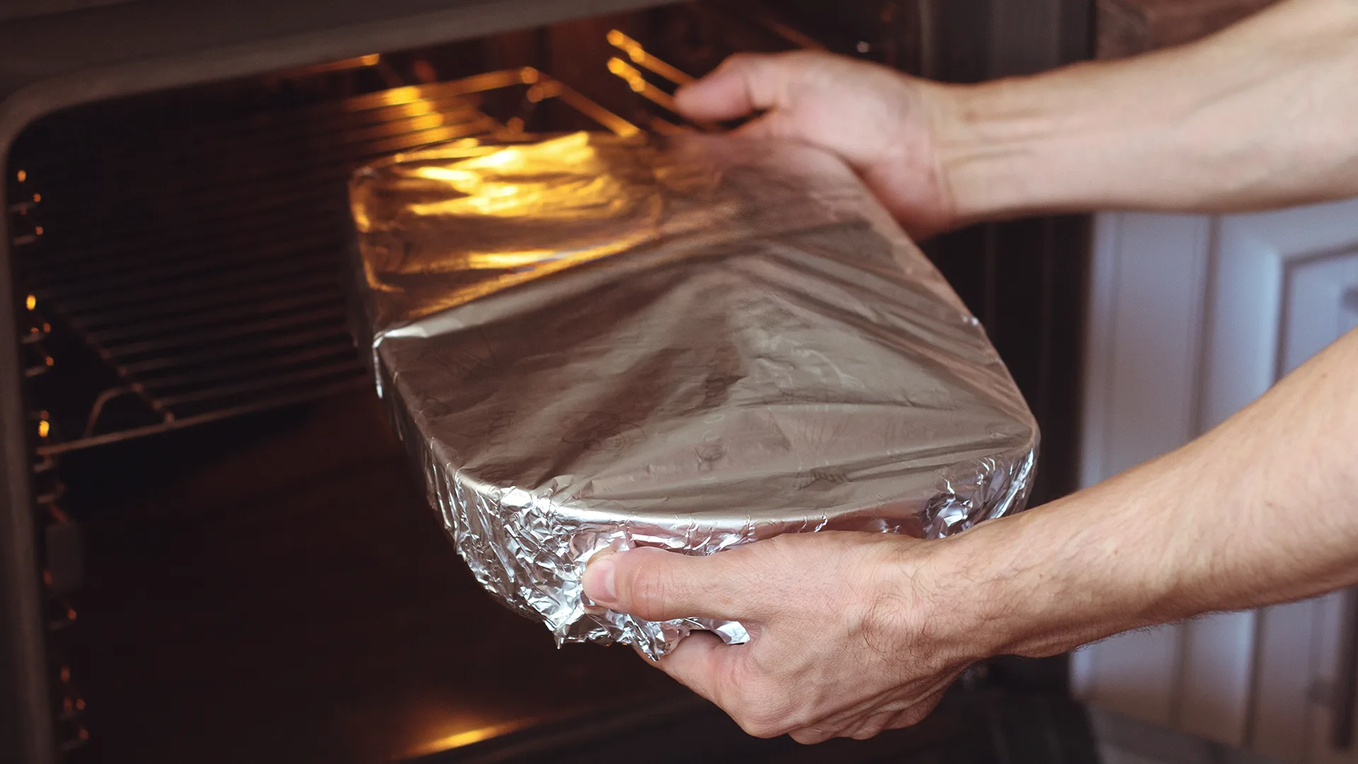 A person places a foil-covered dish into an oven, with the warm glow of the interior light, suggesting food being baked or cooked.