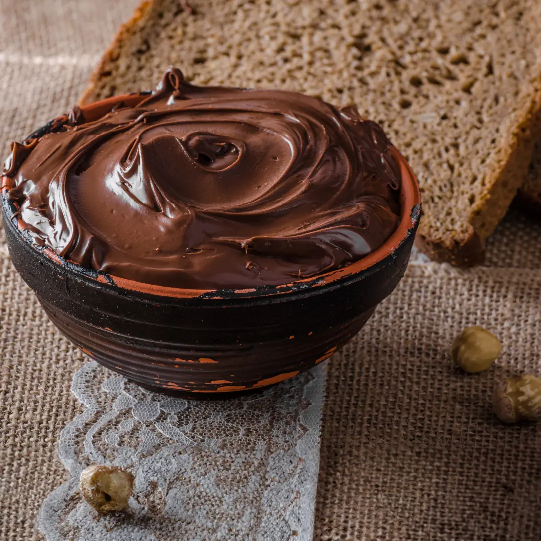 A bowl filled with smooth brown chocolate spread, with slices of brown bread in the background.