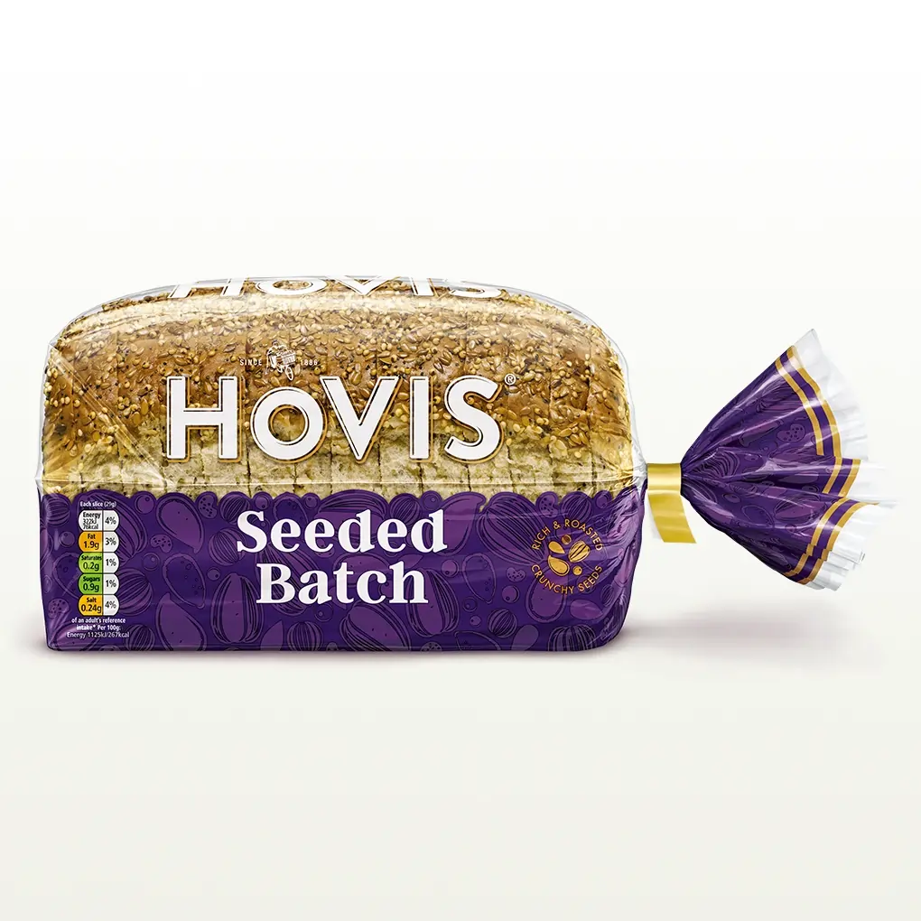 Hovis Seeded Batch bread on a white backdrop with a subtle shadow. The packaging is half transparent and half purple, featuring images of seeds and a label that reads 'Rich & Roasted, Crunchy Seeds.'