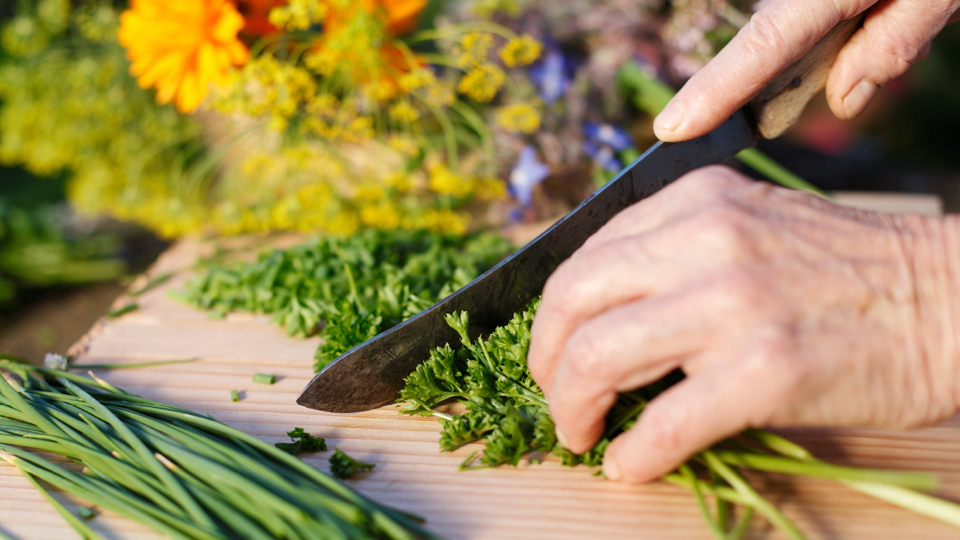 A hand holds down some herbs on a wooden chopping board while the other uses a sharp knife to chop them.