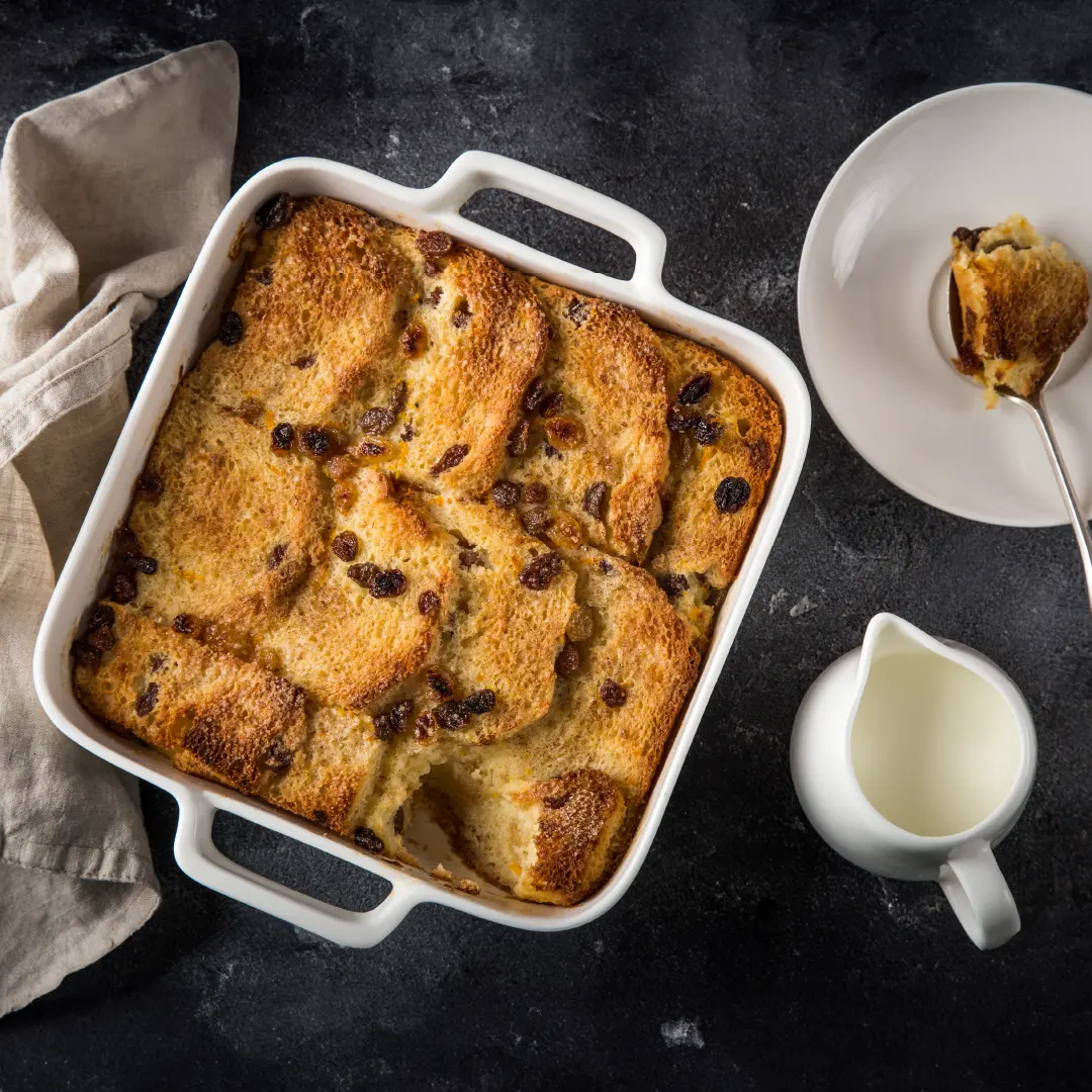 A top down view of a square baking tray with layered toasted Hovis Farmhouse Batch bread topped with raisins. Next to it, a small plate with a spoonful of bread pudding.