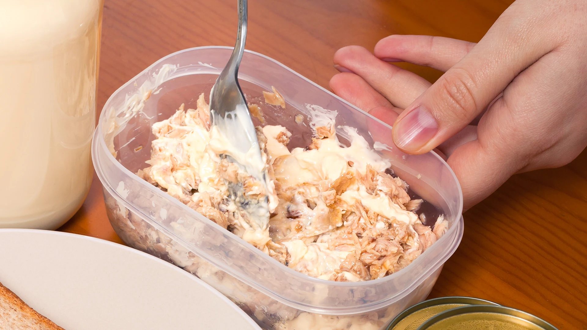 A hand using a metal spoon to mix mayonnaise and tuna in a small rectangular plastic container.