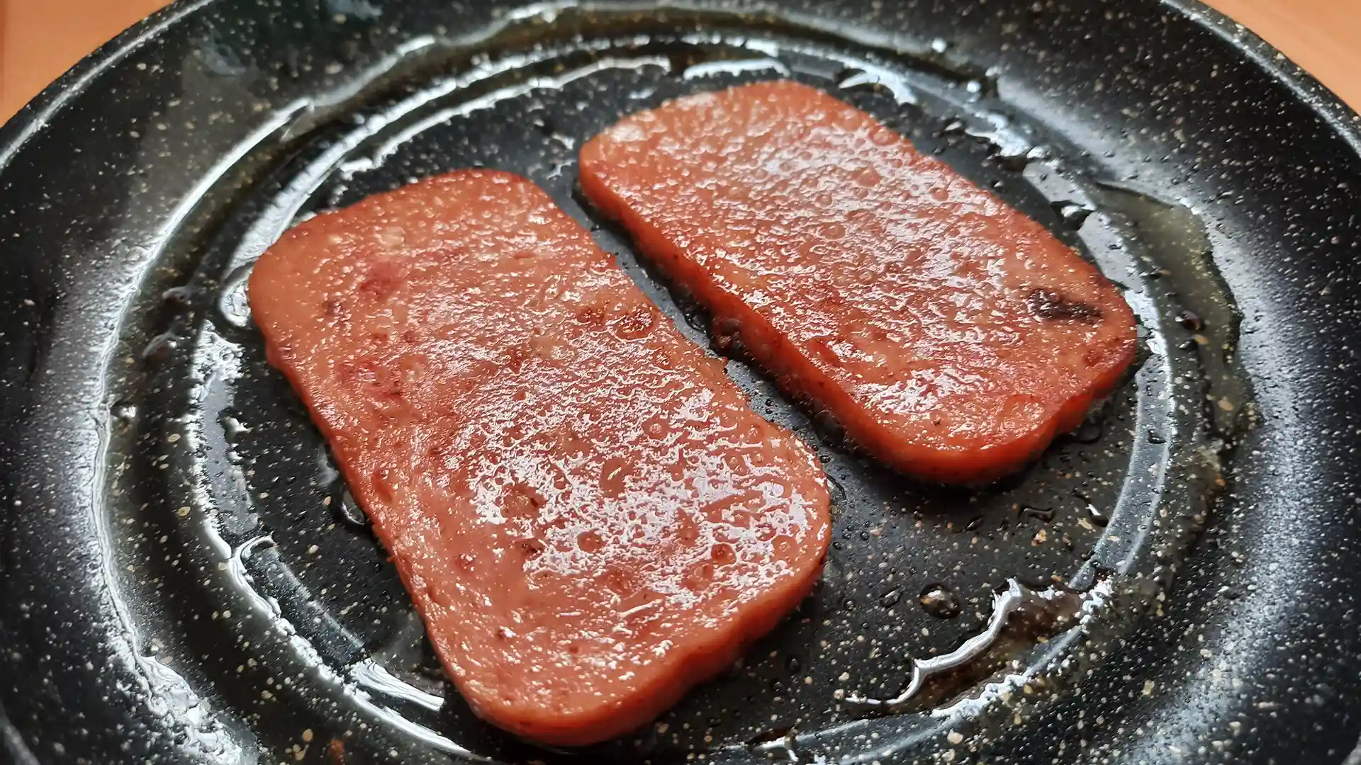 Two crispy slices of spam being fried in a pan.