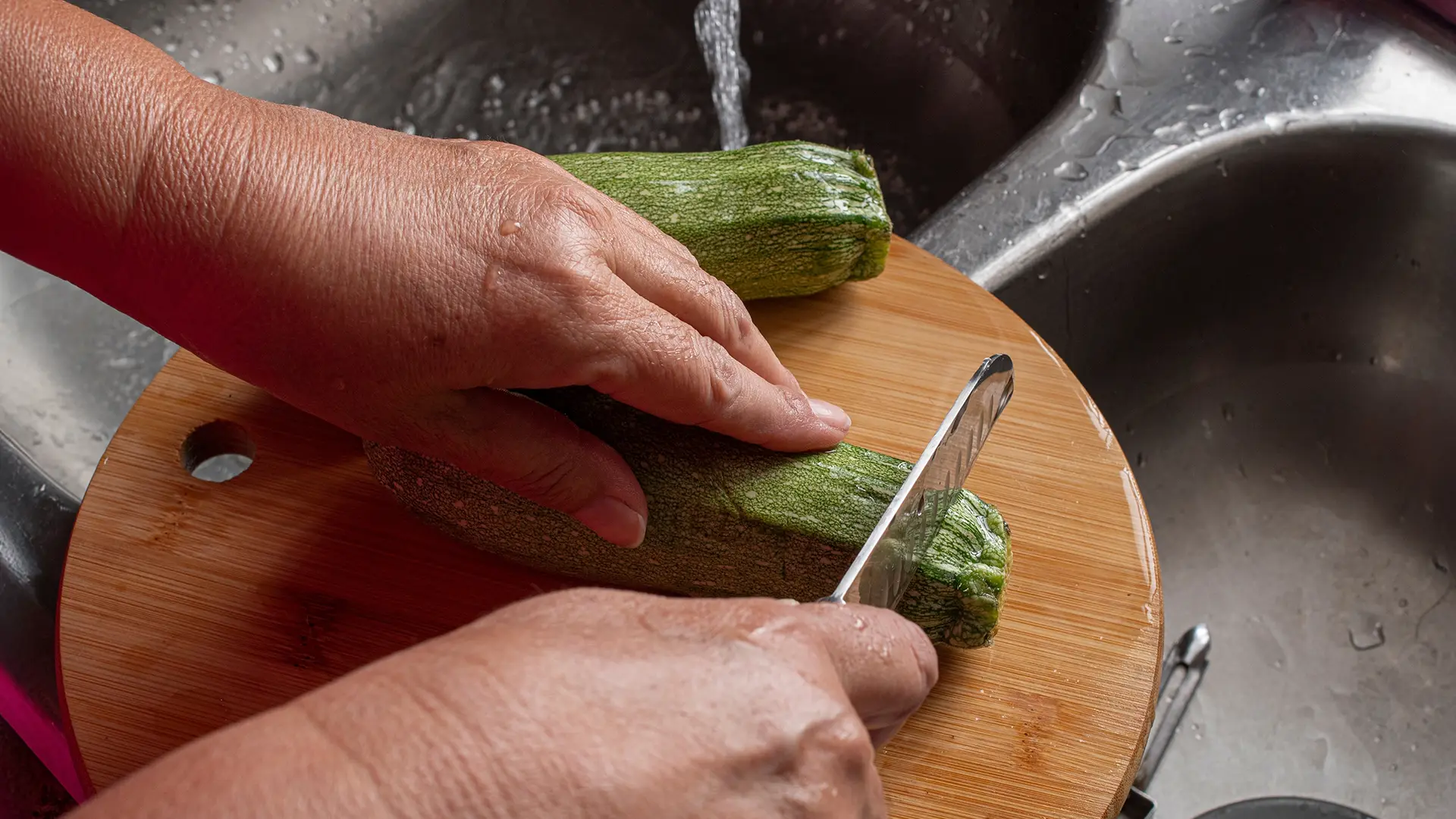 Someone uses a knife to cut a courgette on a wooden chopping board, near a kitchen sink. An extra courgette can also be seen on the chopping board.